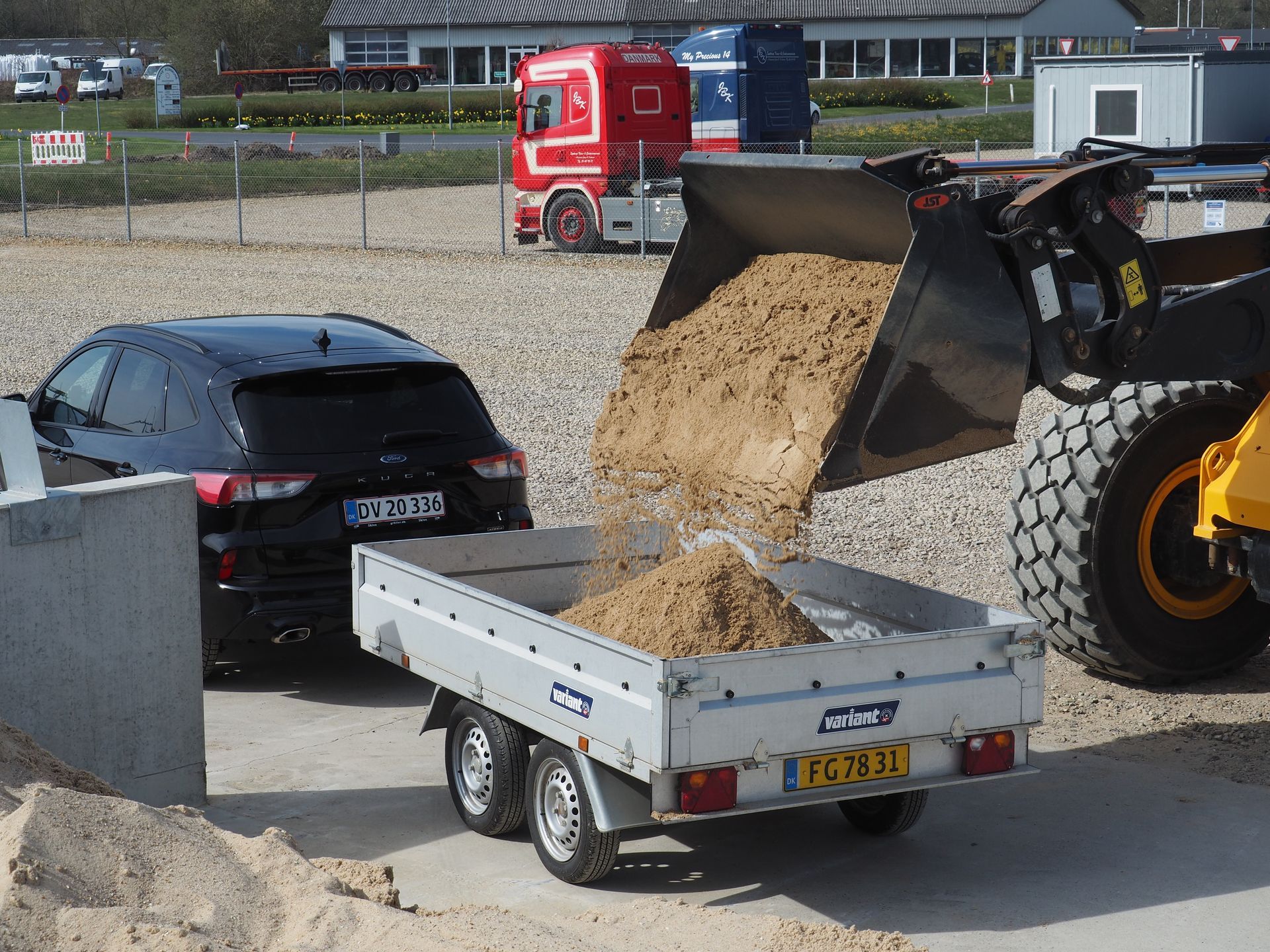 En frontlæsser tømmer sand i en trailer ved siden af ​​en sort bil. Lastbiler er i baggrunden.