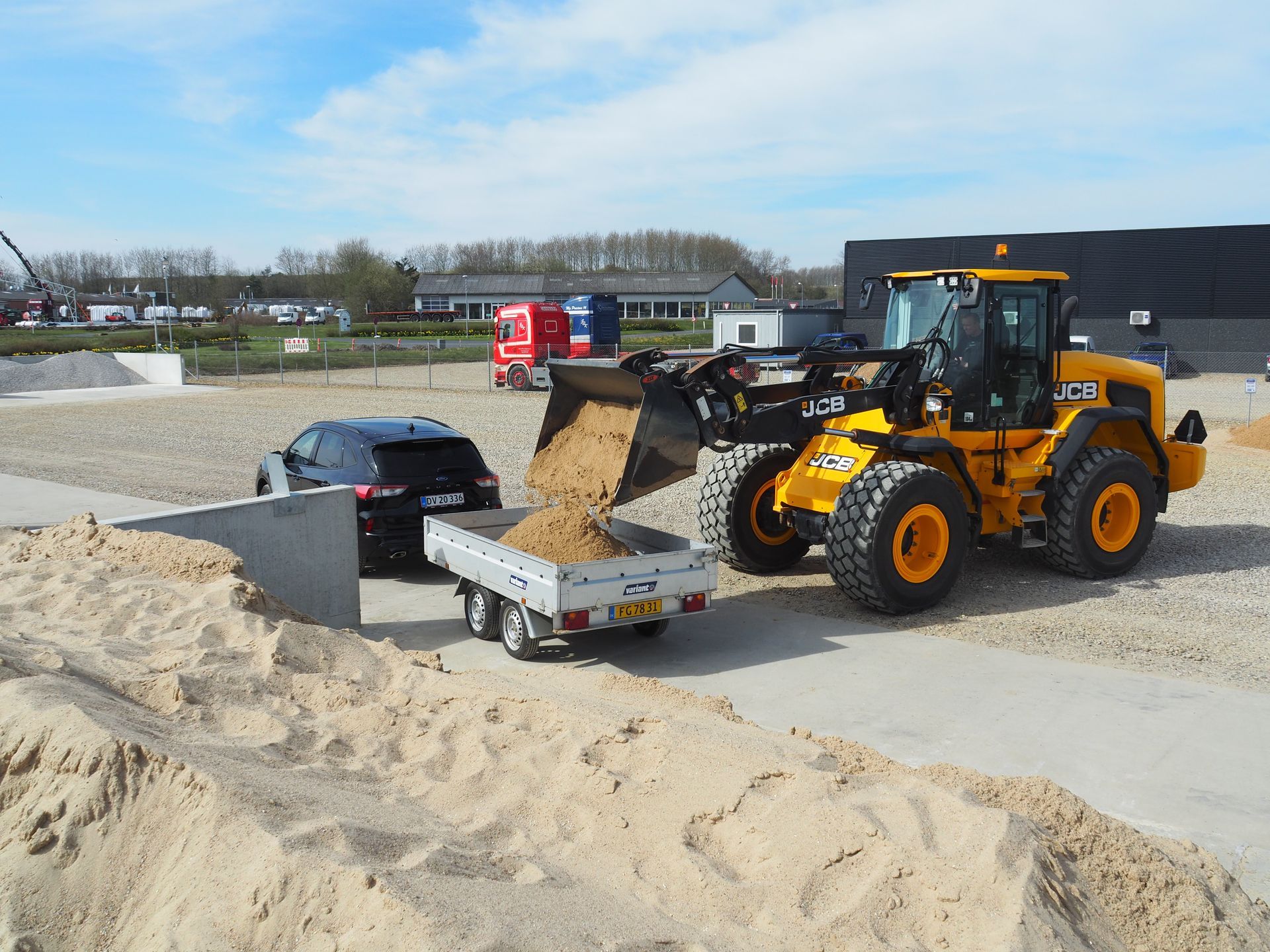 En sølvfarvet trailer ved siden af ​​en bunke sand med en betonvæg i baggrunden på en udendørs byggeplads.