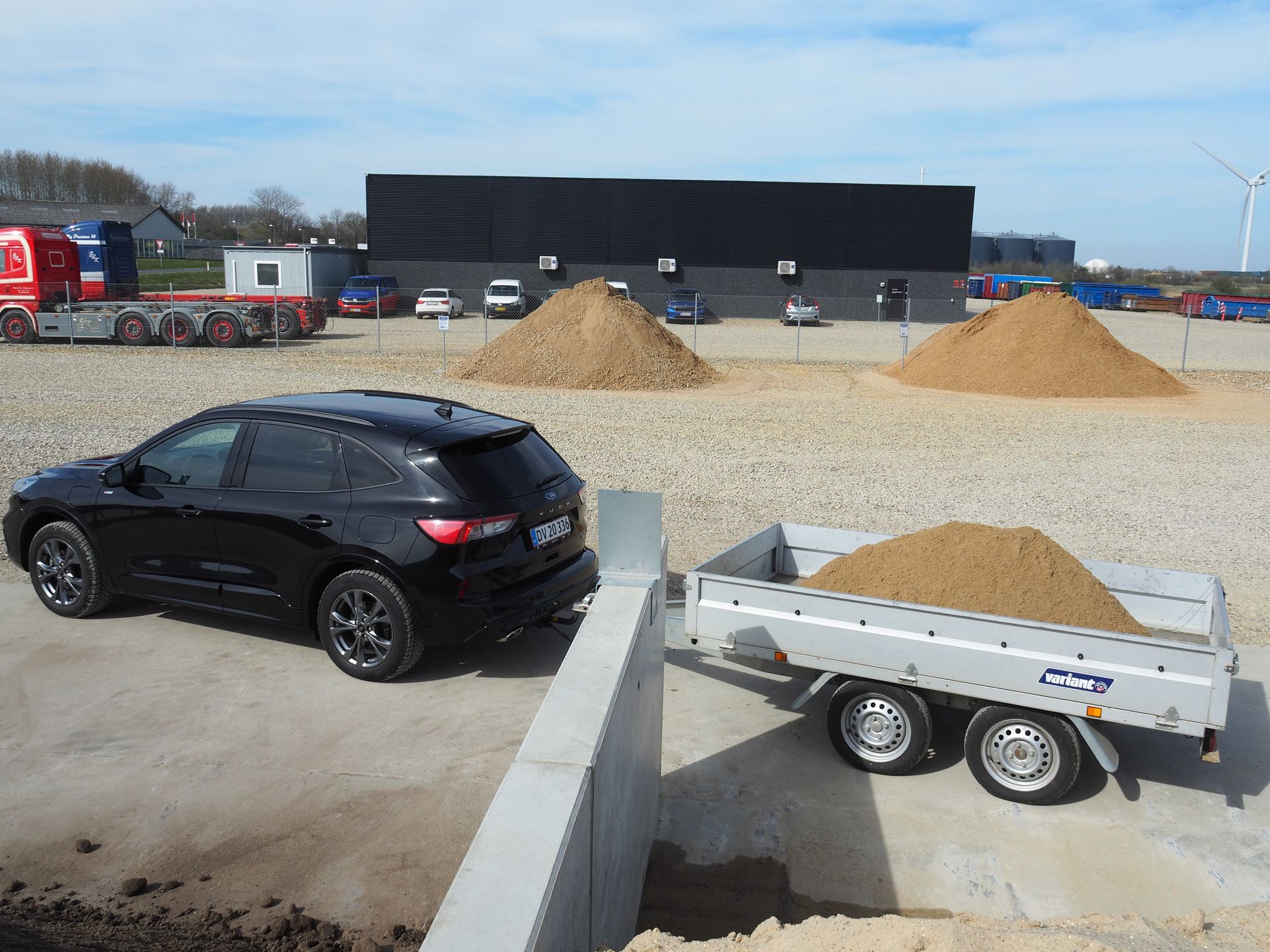 En sølvfarvet trailer ved siden af ​​en bunke sand med en betonvæg i baggrunden på en udendørs byggeplads.