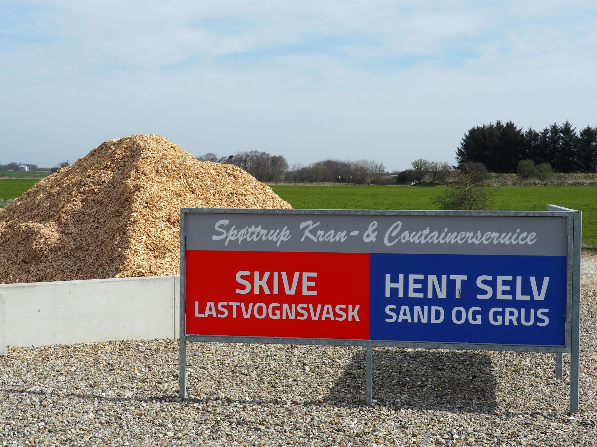 Skilt ved en affaldsplads med træflis og grus, ved siden af ​​en parkeret bil.