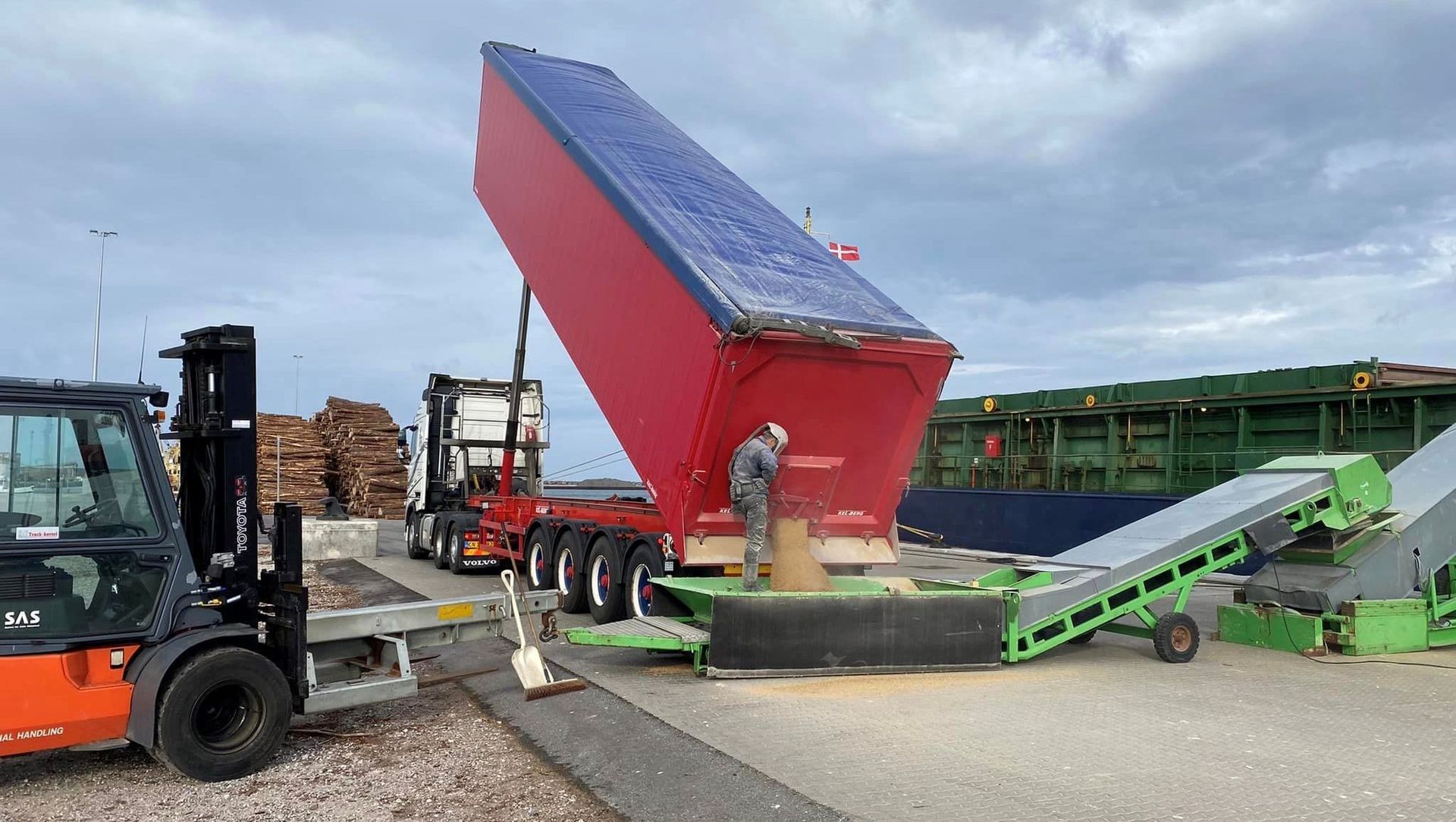 En rød trailer tømmer materiale på et grønt transportbånd; en person overvåger processen.