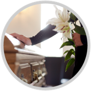 Person placing white lilies on a polished wooden casket in a bright funeral setting