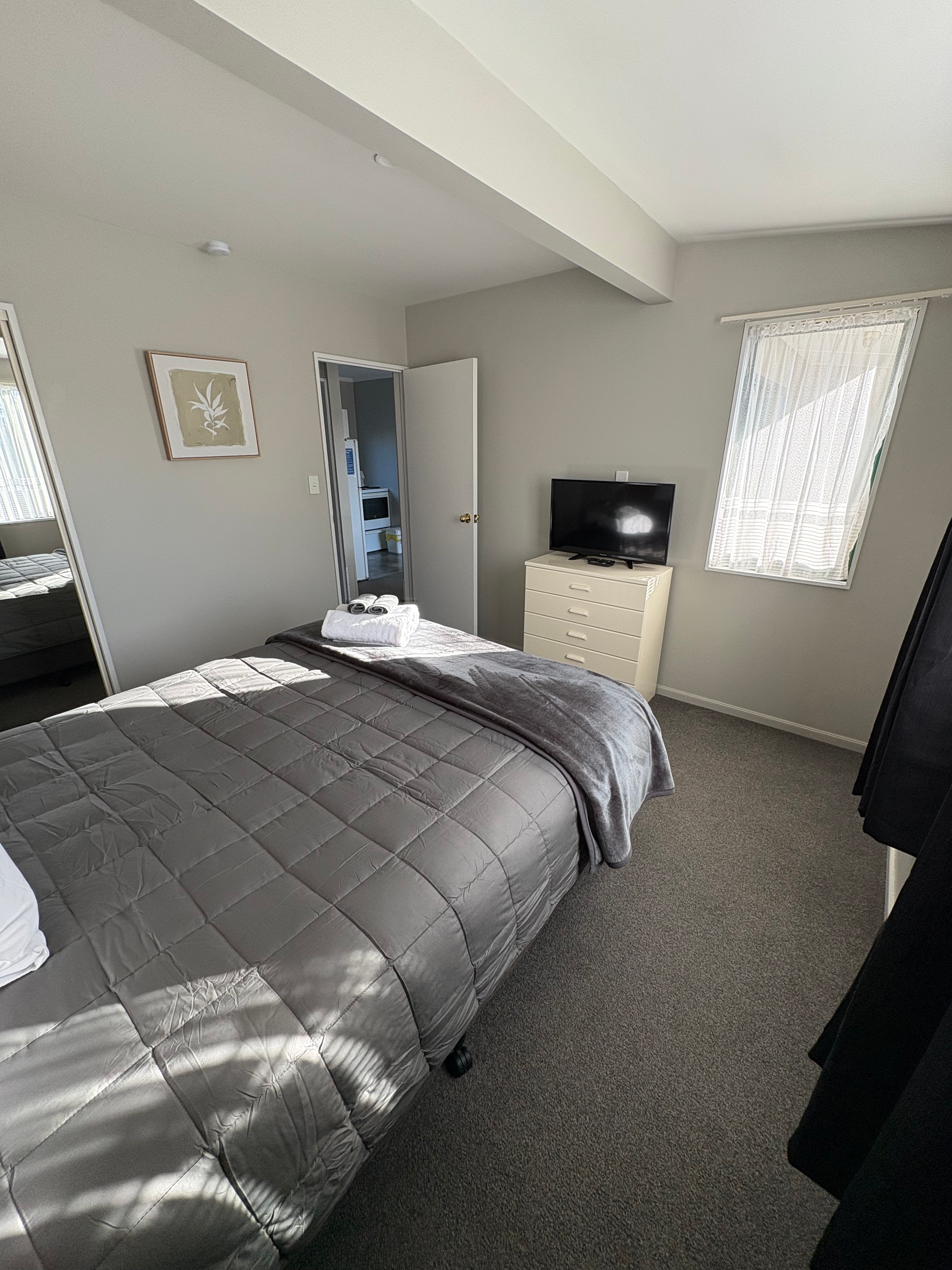 Bedroom with gray bedspread, open closet, small dresser, and window with sheer curtains.