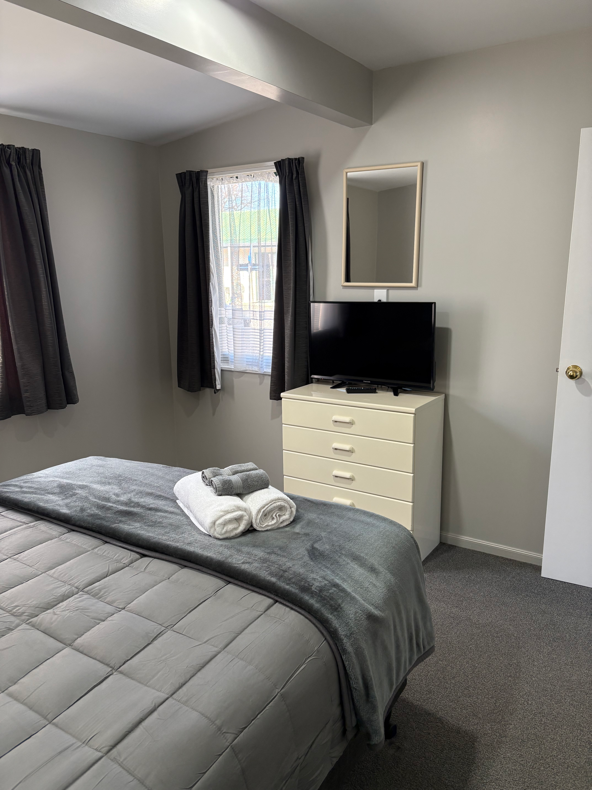 Bedroom with gray bedspread, folded towels, dresser, mirror, TV, and window with dark curtains.