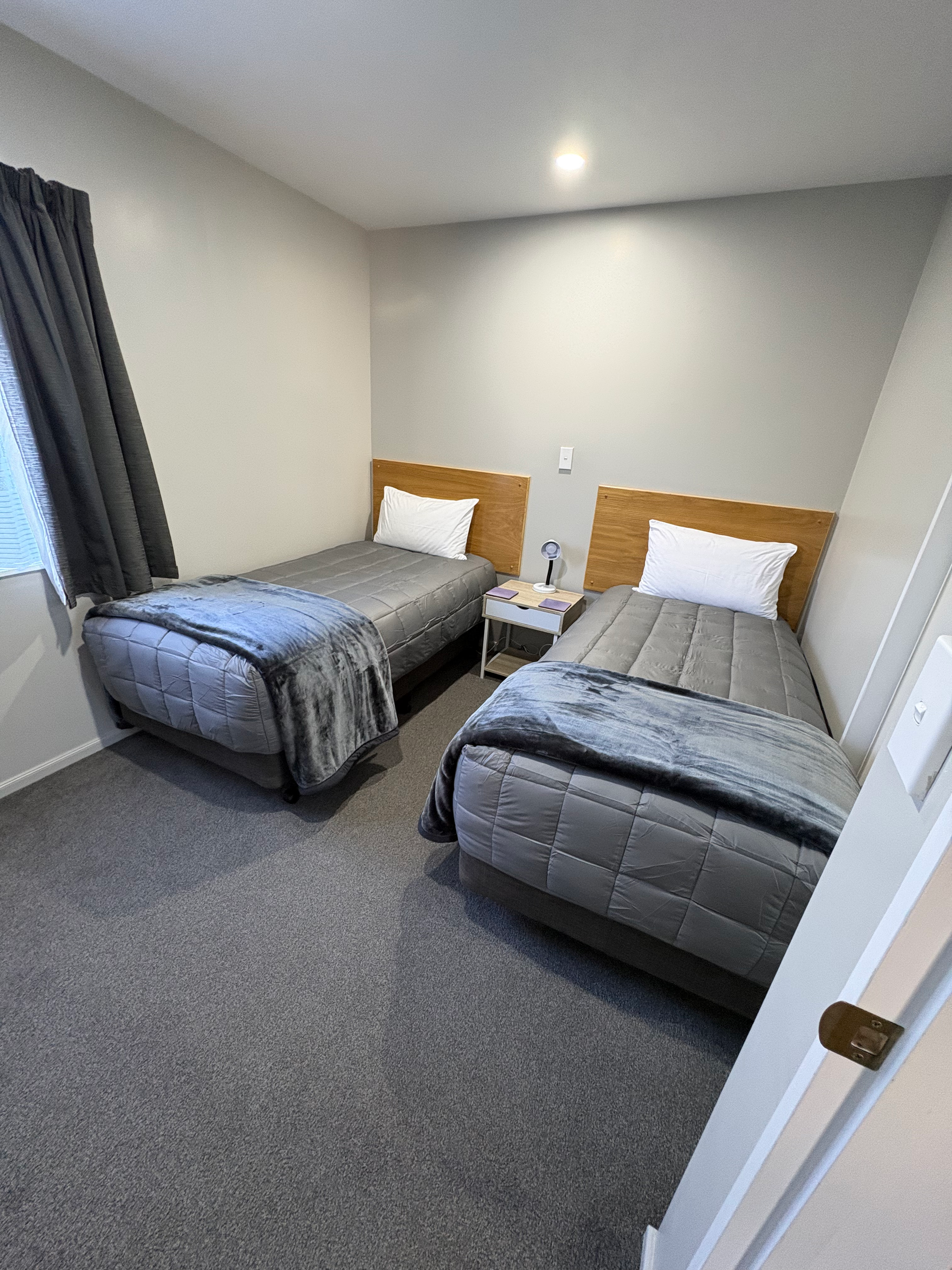 Small bedroom with two twin beds, gray bedding, white walls, and a window with dark curtains.