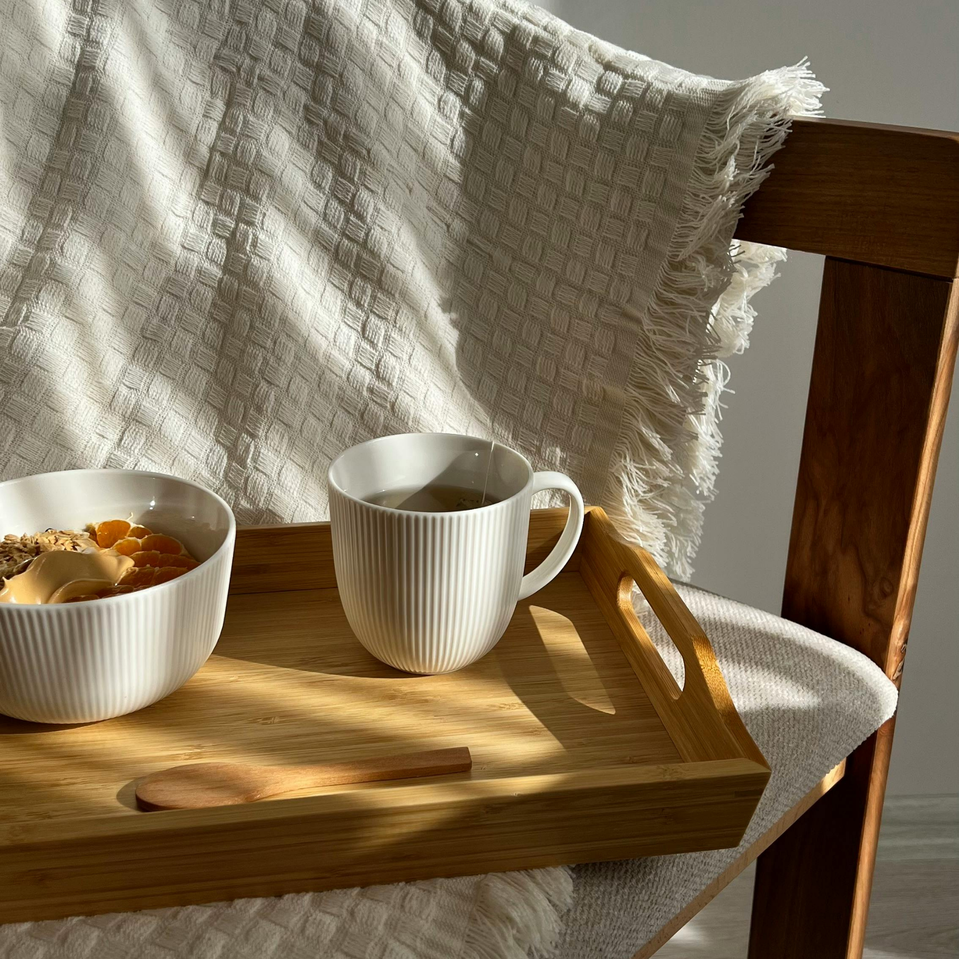 Breakfast tray with a white mug, a bowl of cereal, and a wooden spoon on a sunlit bed