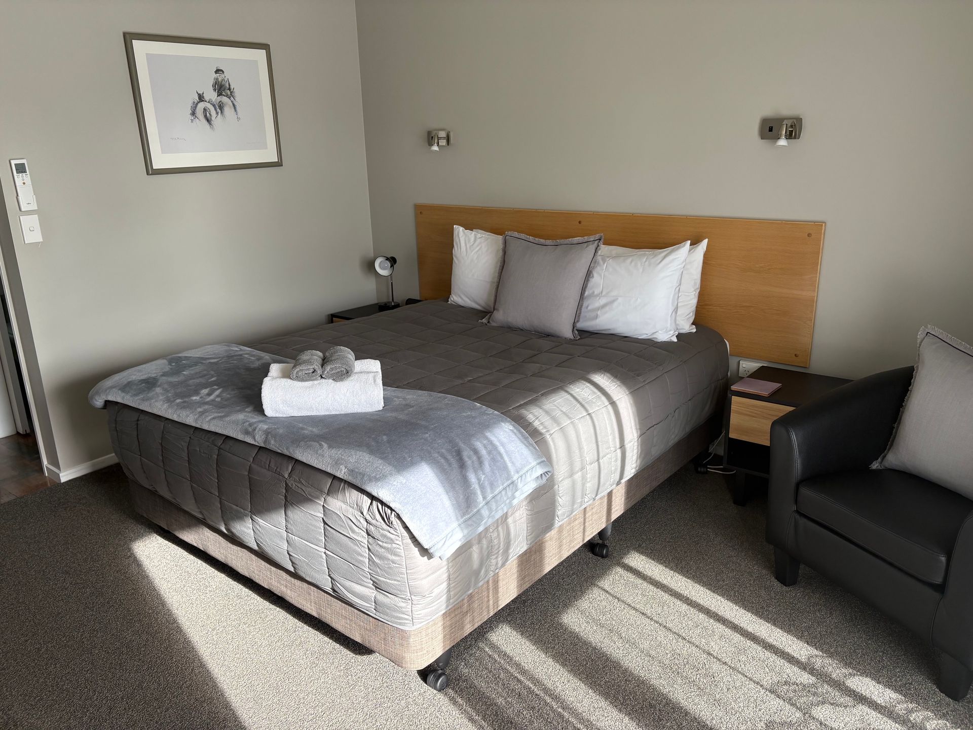 Neatly made bed with gray bedding, bedside table, armchair, and sunlight on a carpeted hotel room floor.
