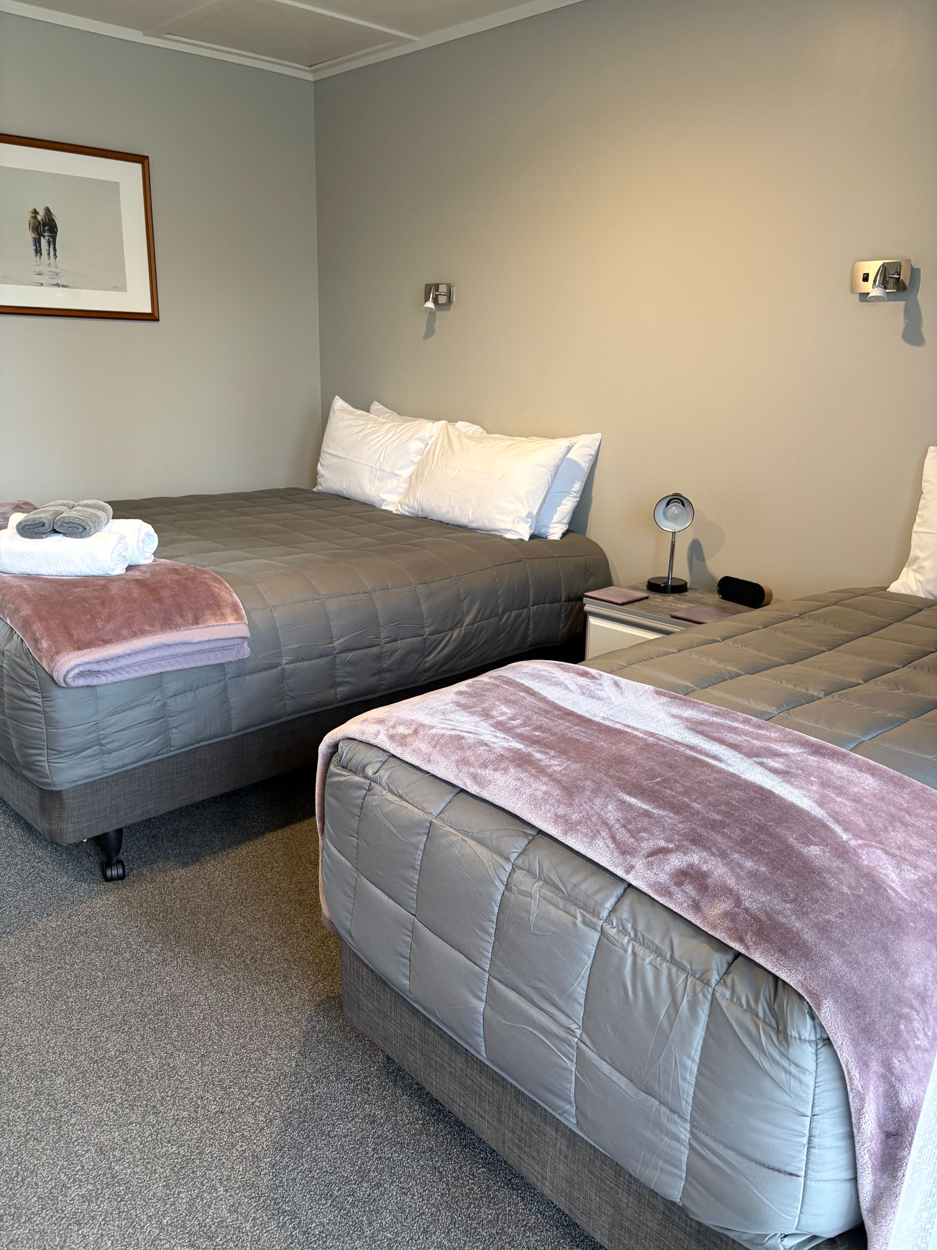 Two neatly made twin beds in a gray hotel room with pink blankets and wall lamps