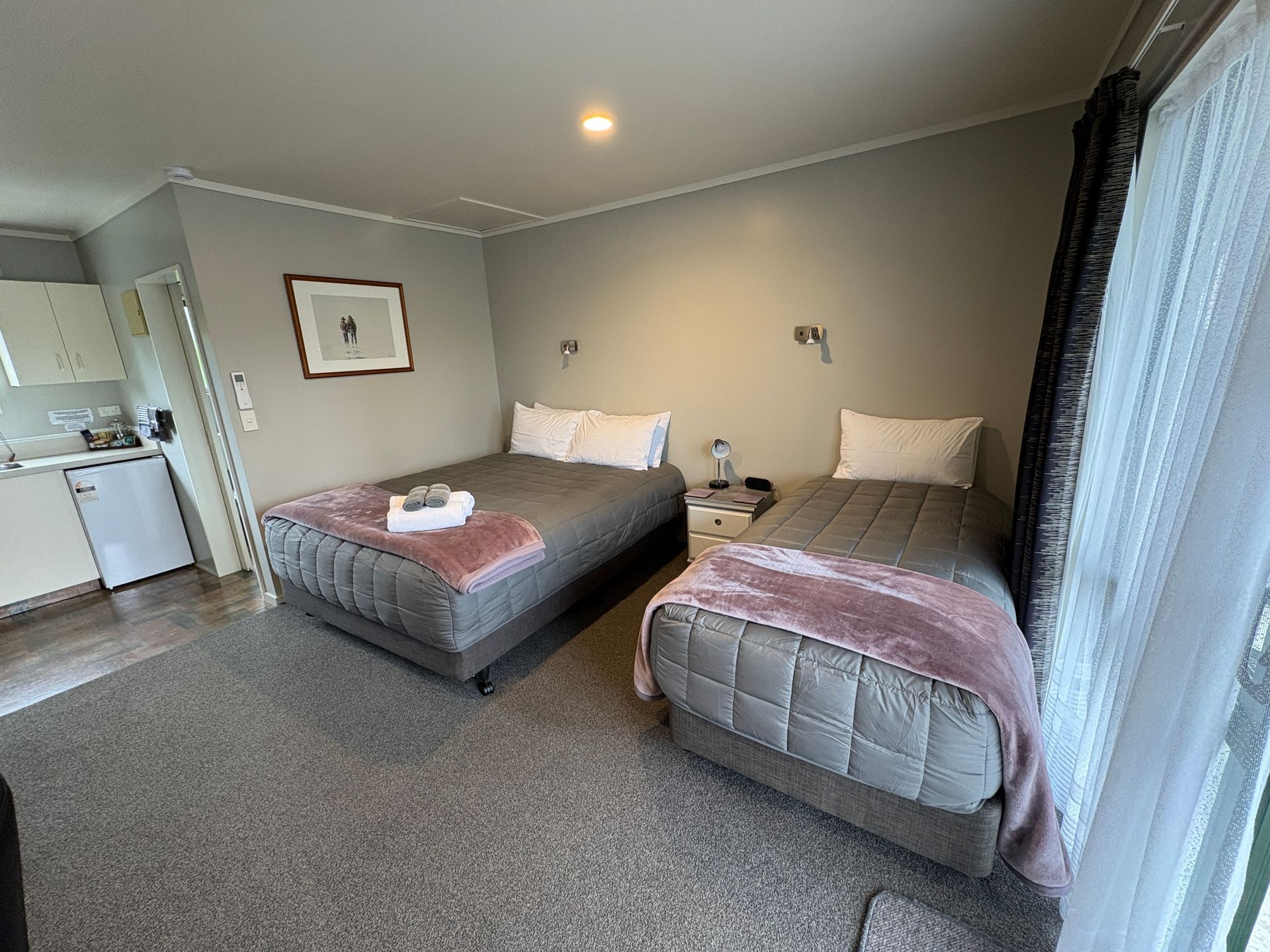 Small hotel room with two beds, gray carpet, and a kitchenette by the door.