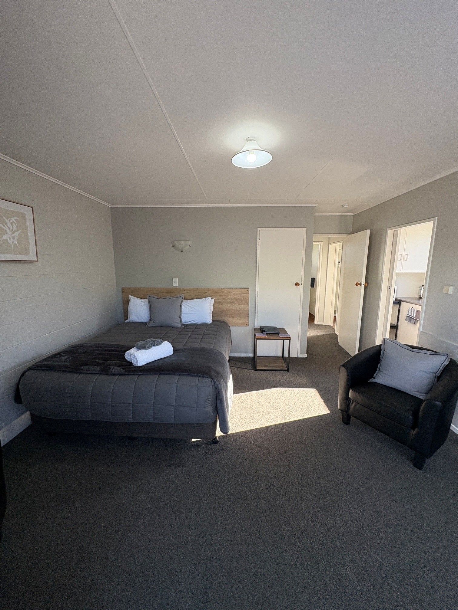 Neat bedroom with a bed, chair, and open doorway, lit by a ceiling light and sunlight.