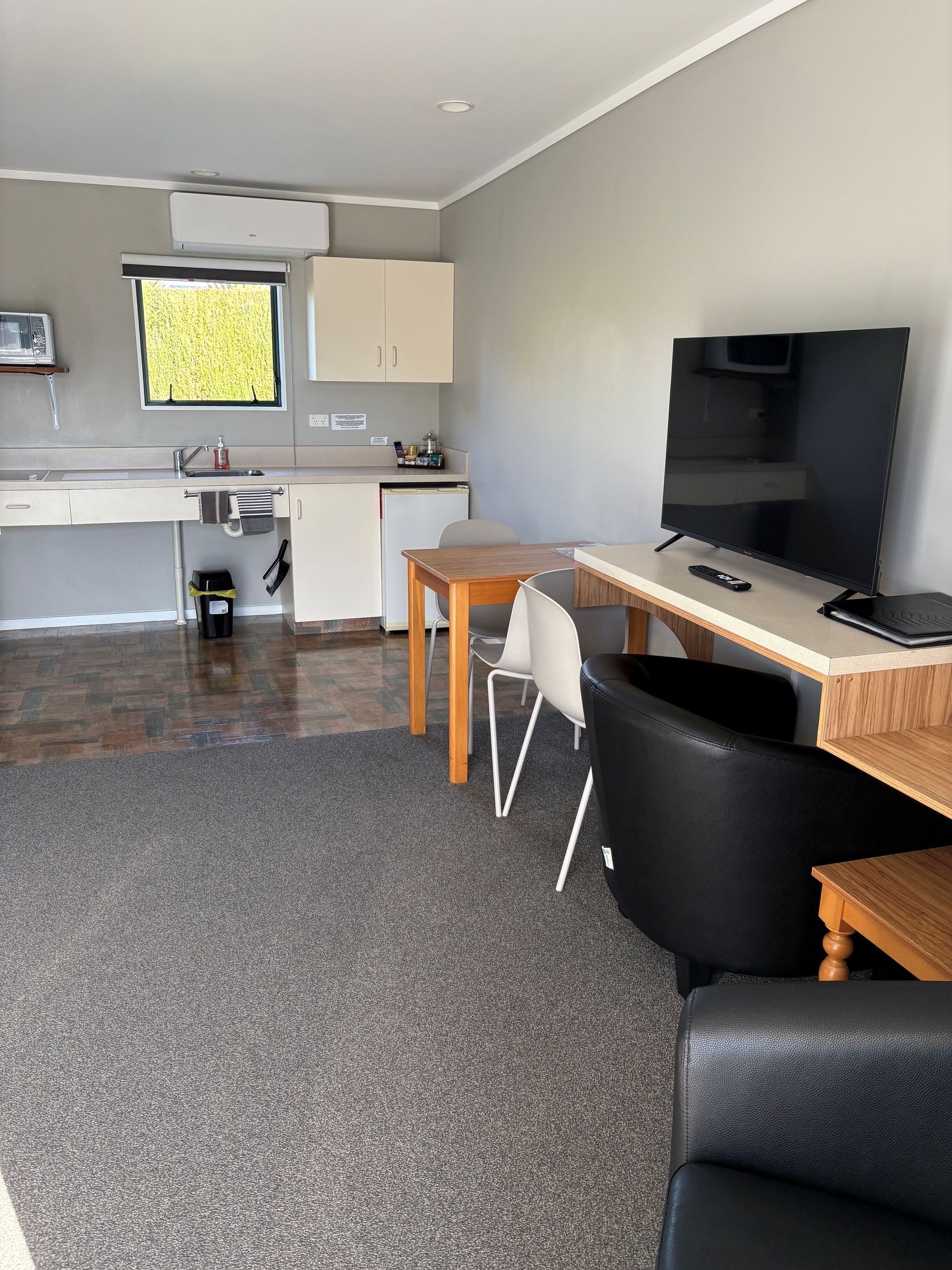 Small studio room with kitchenette, desk, TV, and black chairs on a speckled carpet.