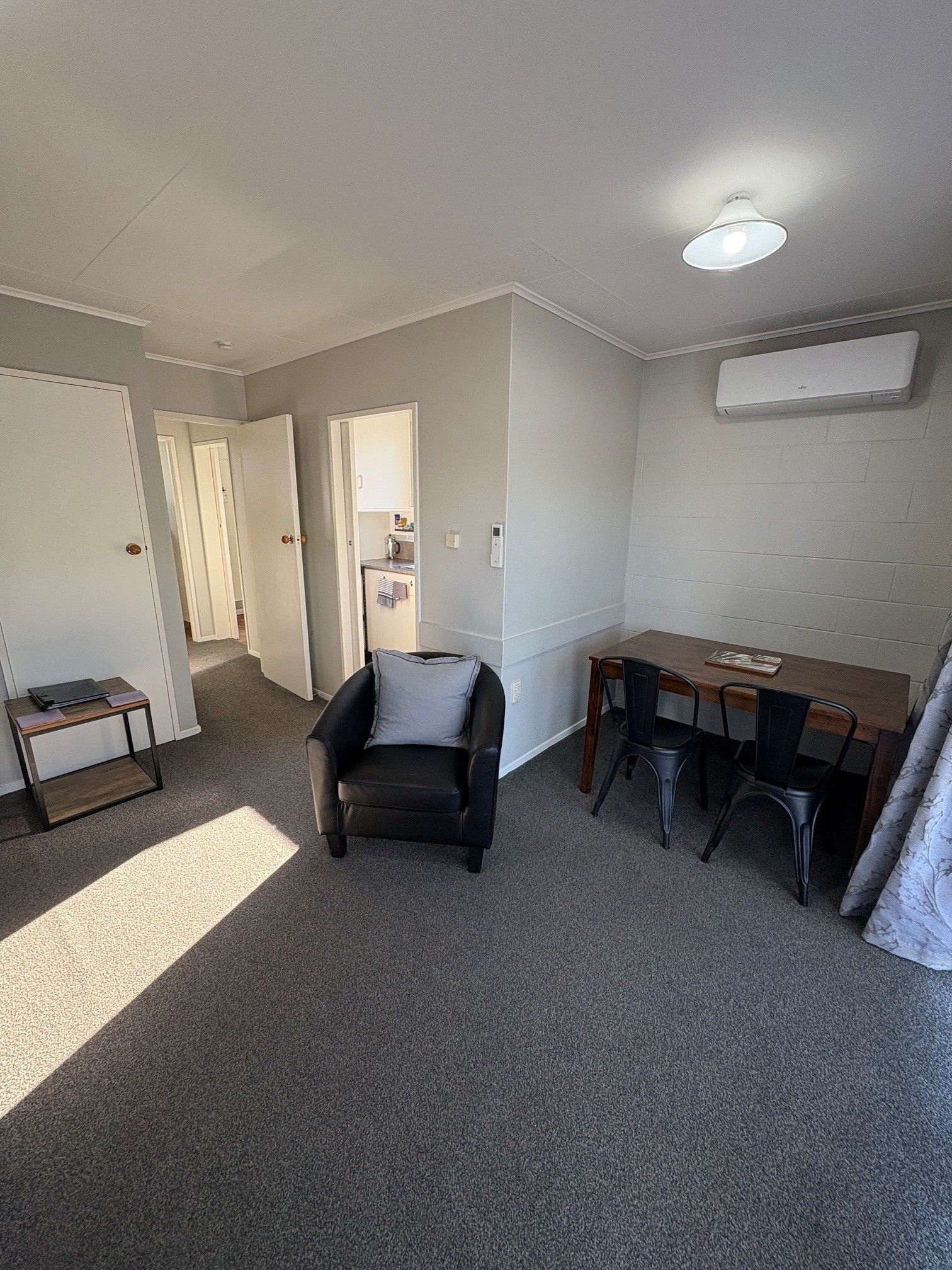 Small carpeted room with armchair, desk and chairs, wall-mounted heater, and open doorway to a kitchen.