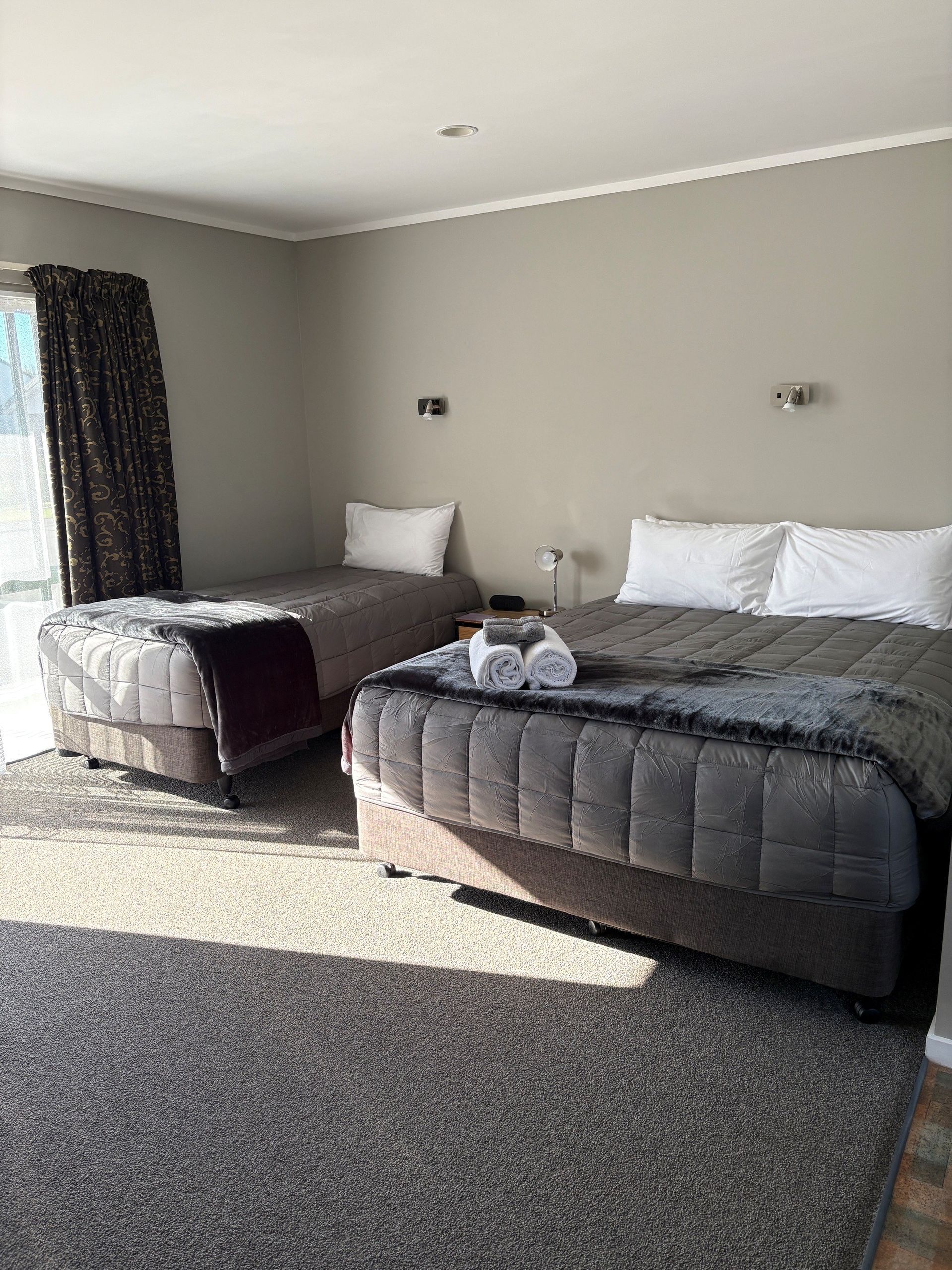 Hotel room with two neatly made beds, gray carpet, and sunlight from a window.