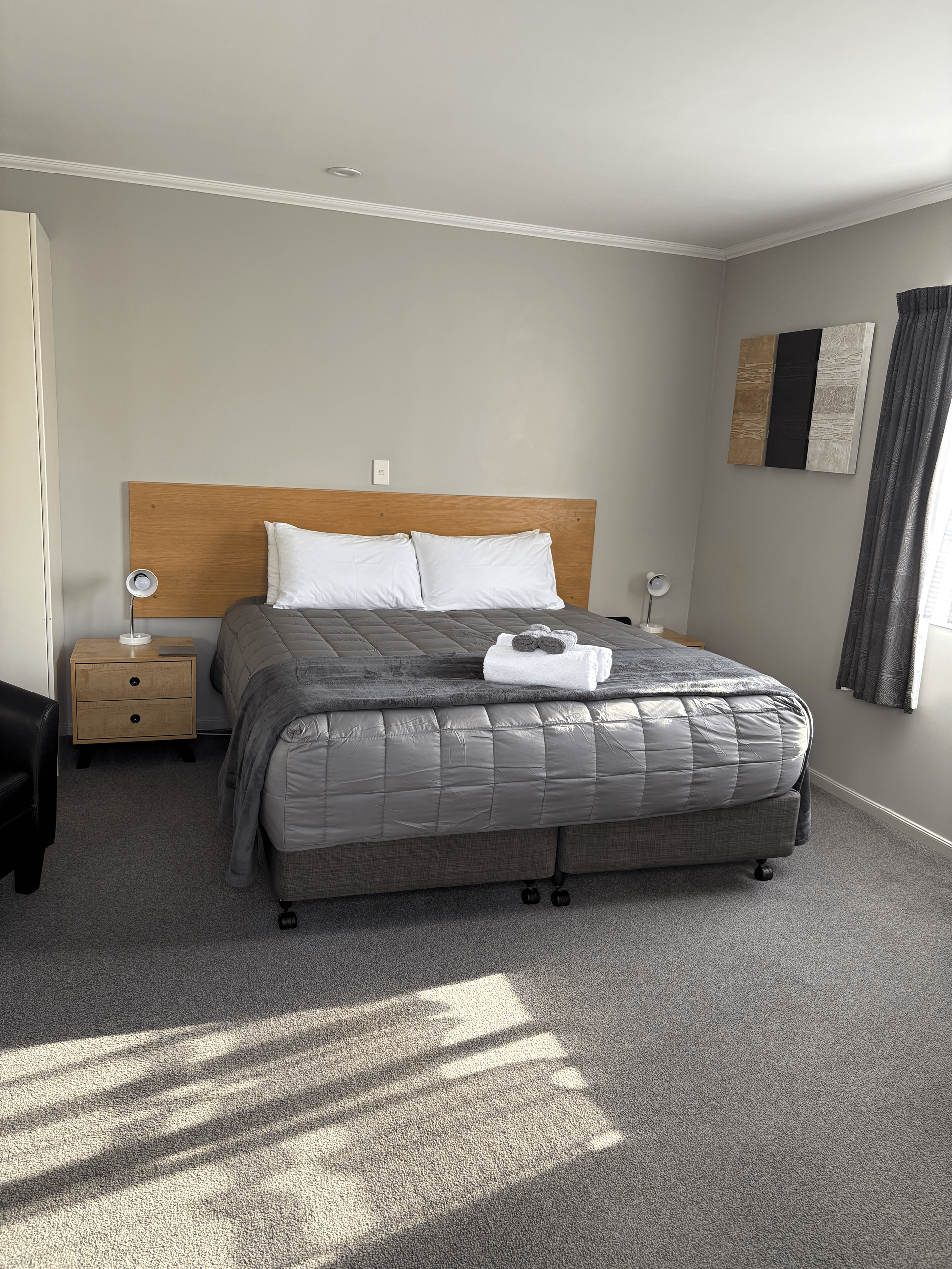 Bedroom with a gray bed, wooden headboard, nightstand, wall art, and sunlight on carpet.