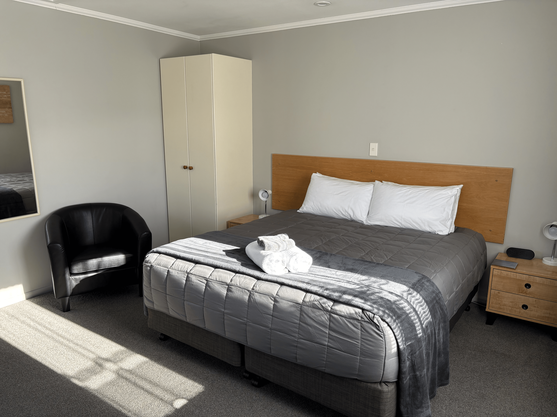 Bright bedroom with gray bed, white pillows, black chair, wardrobe, and sunlight on carpet.