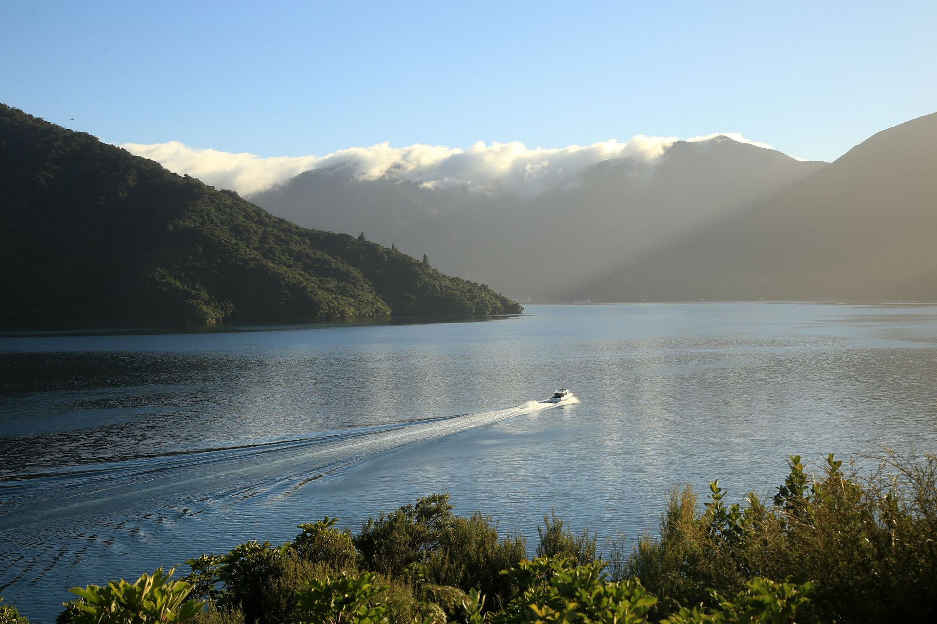 Lake with a small speedboat, forested hills, and sunlit mountains in the distance