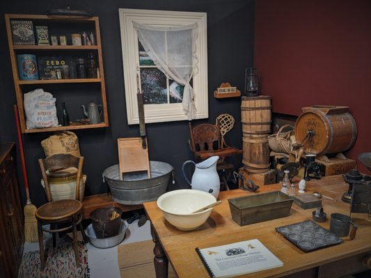 Old-fashioned kitchen display with wooden shelves, metal tubs, crockery, and antique tools
