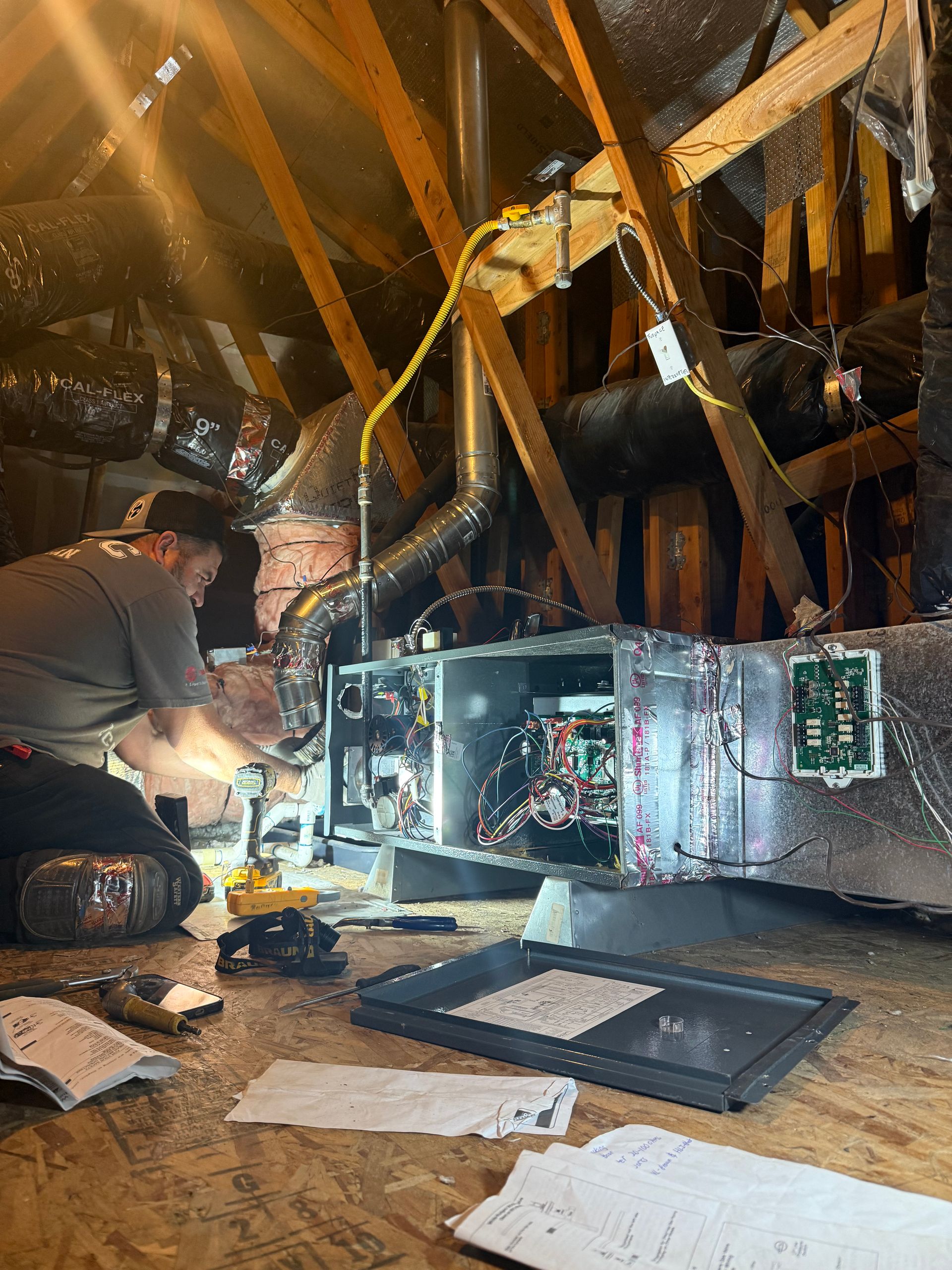 A person in a jumpsuit and hard hat services an air conditioning unit indoors, standing on a ladder.