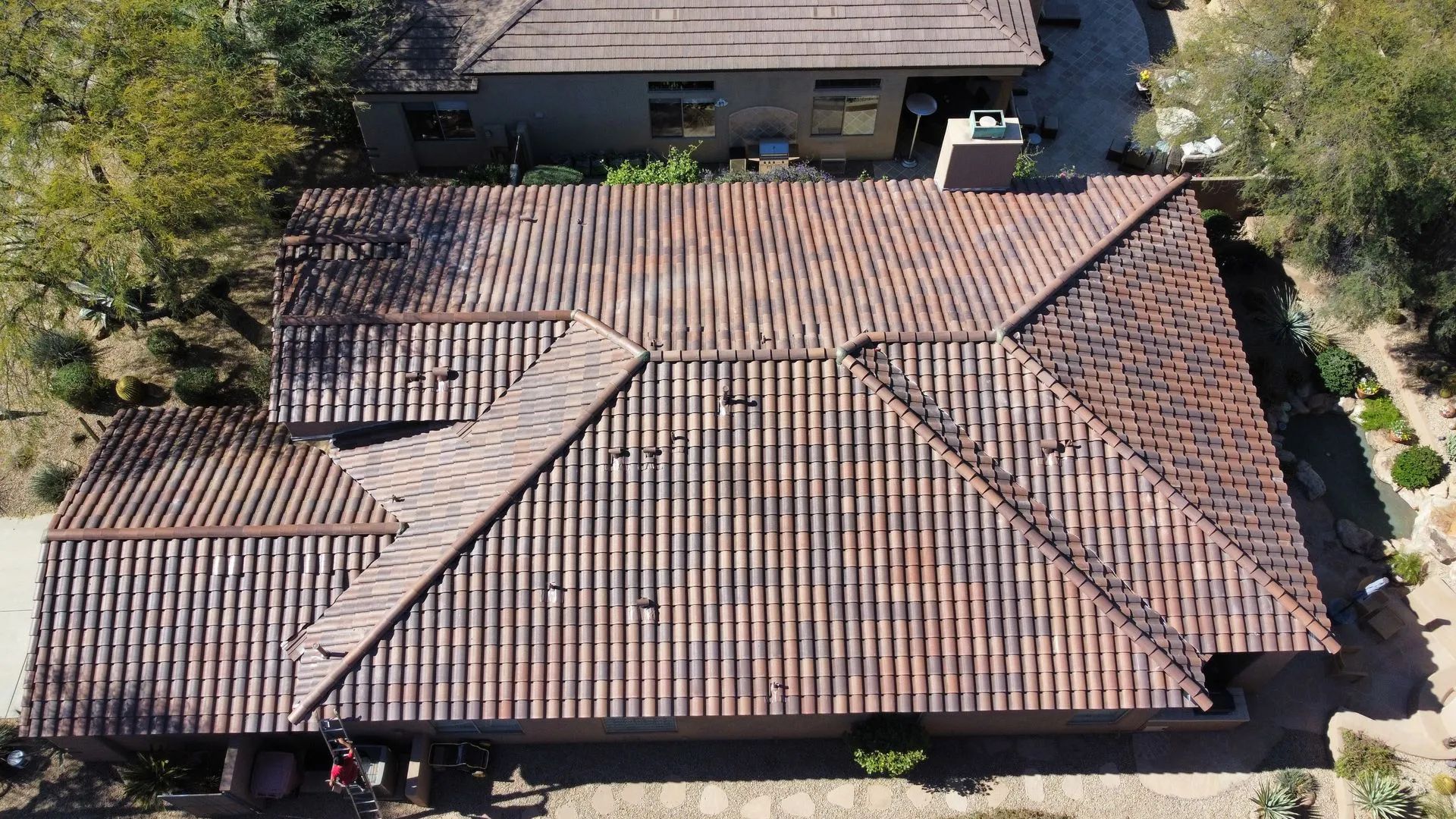 Aerial view of a tan-tiled suburban house roof with multiple gables and surrounding trees.