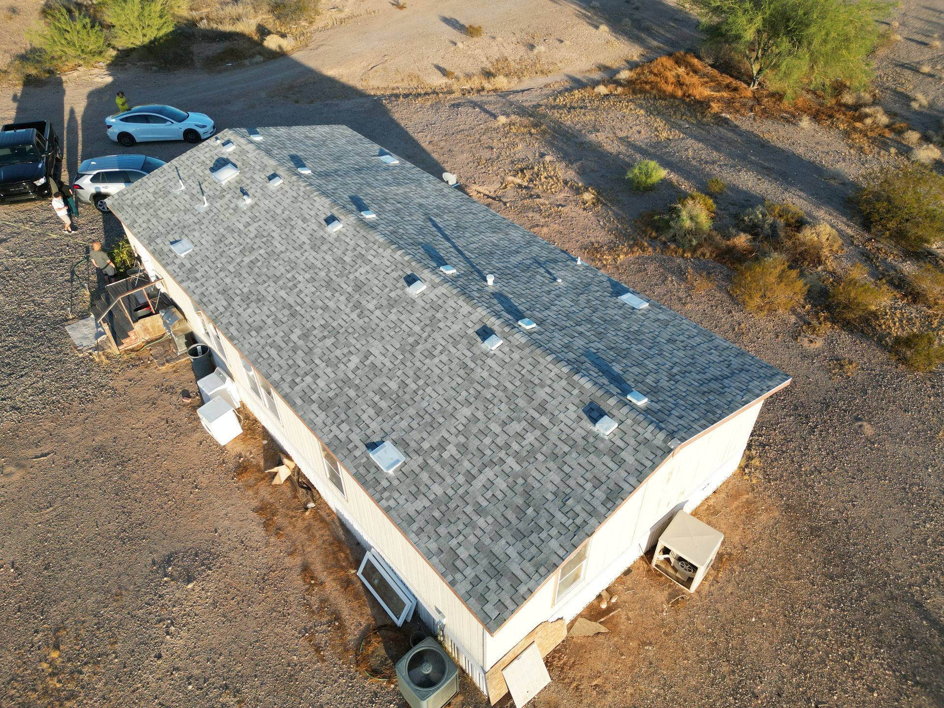 Aerial view of a small white building with a gray roof in a rocky desert lot, with a blue car nearby.