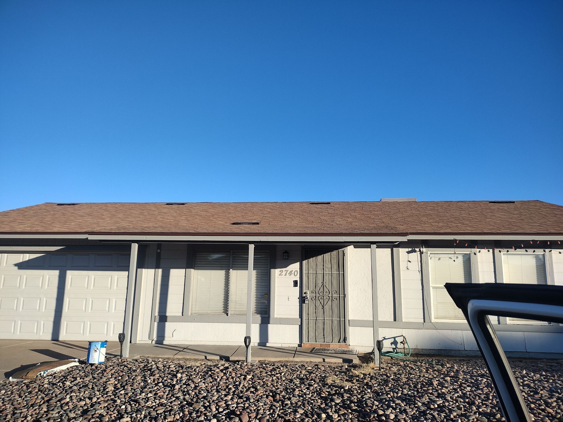 Gray house with brown roof against a clear blue sky. Rocky ground in front. Car door open.
