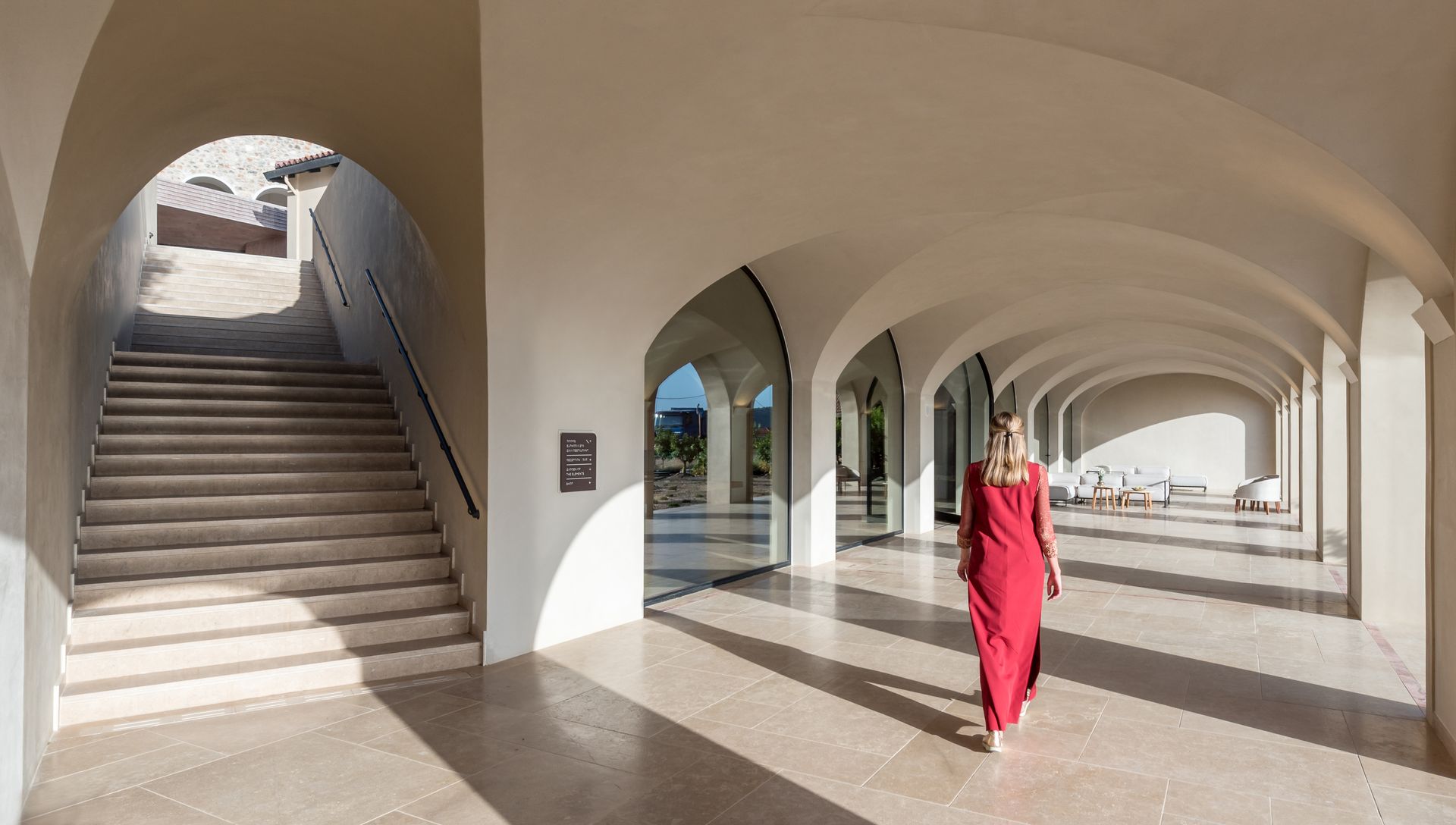 Une femme en robe rouge marche dans un couloir avec des arches et des escaliers.