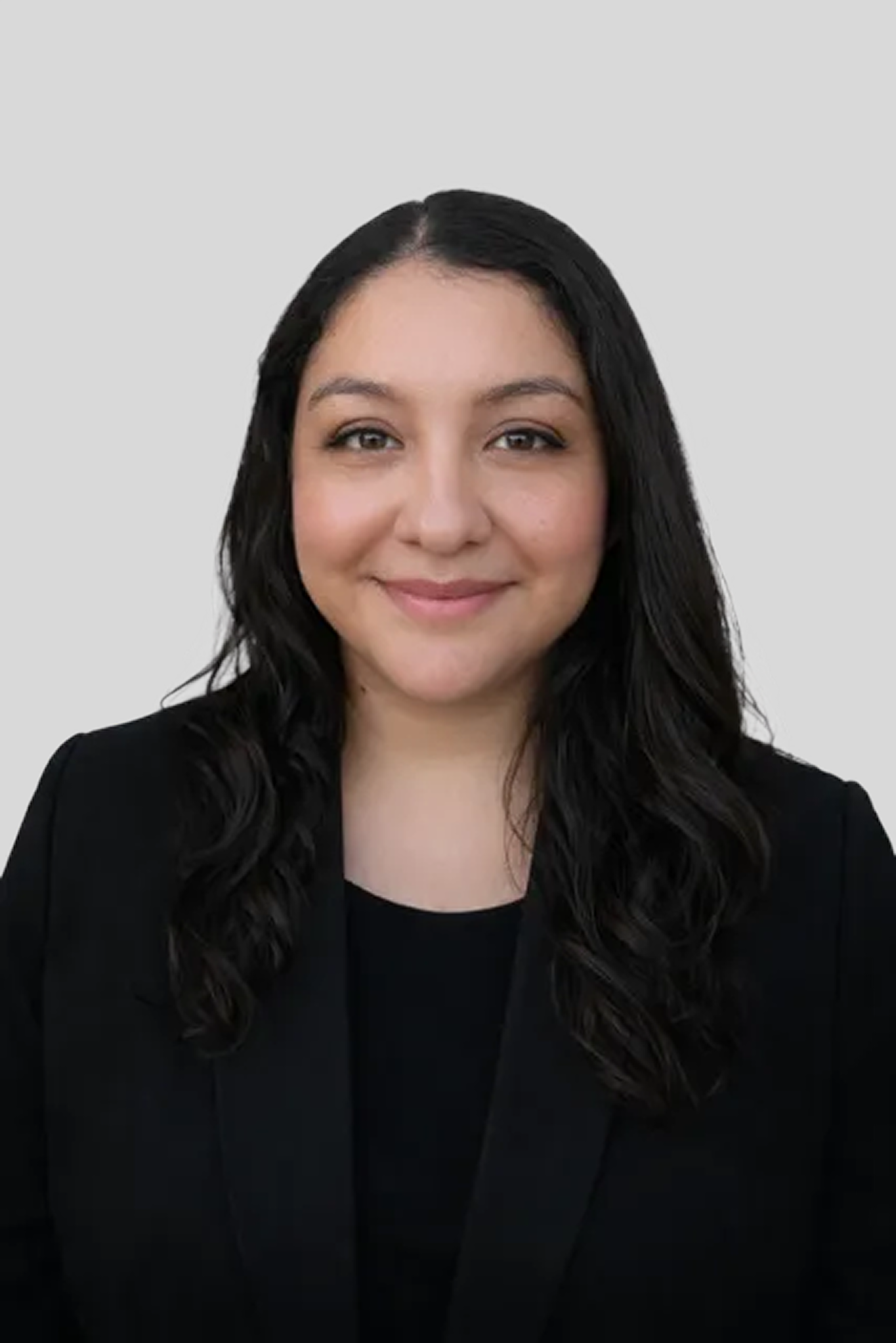 Studio portrait of a smiling person with long dark hair, wearing a black blazer against a light gray background