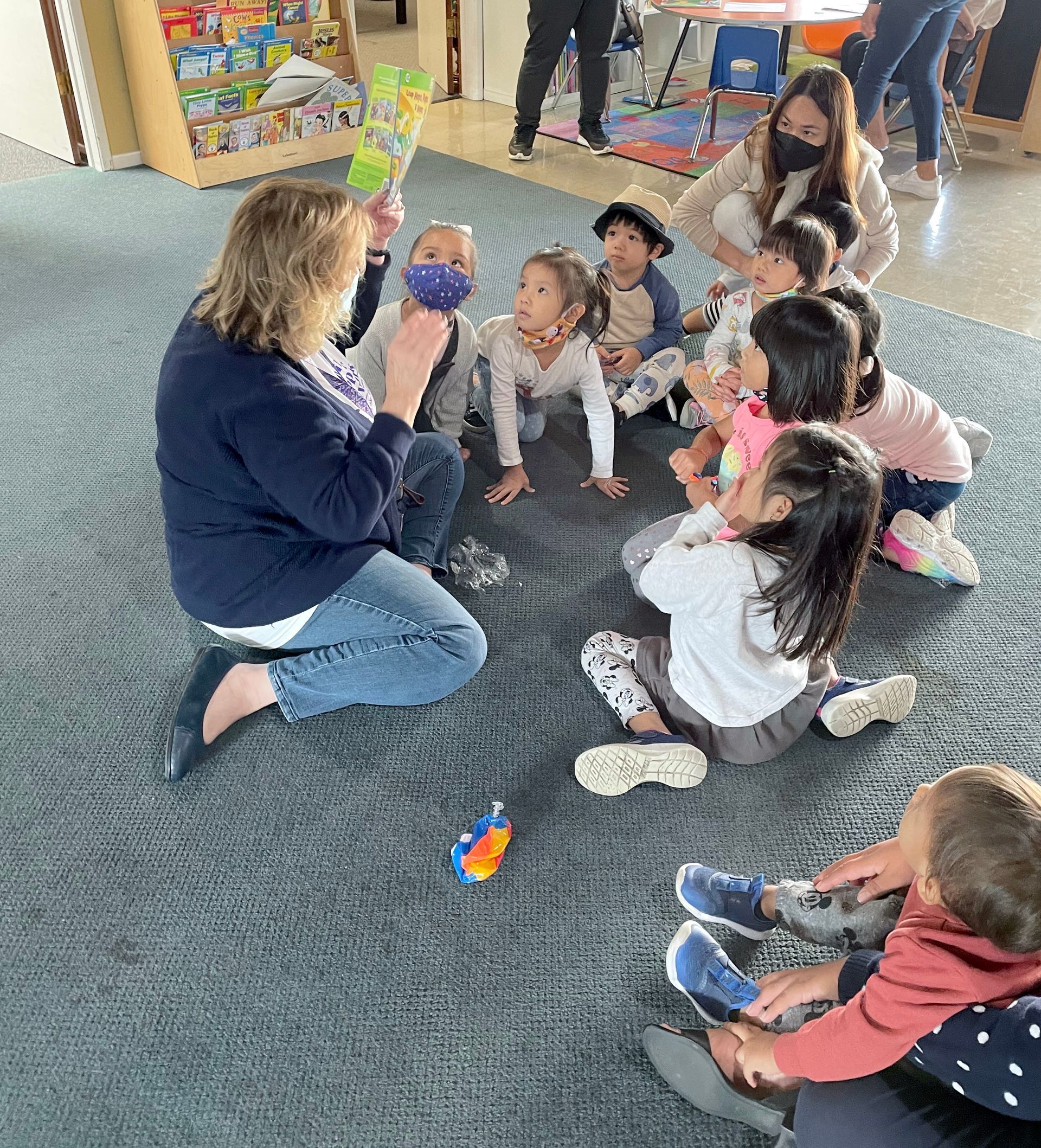 Teacher kneels on carpet, leading a small circle time with children seated around her in a classroom.