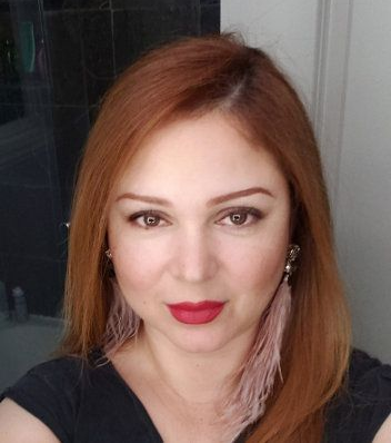 Close-up selfie of a woman with auburn hair, red lipstick, and a black top against a light background