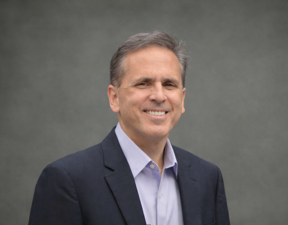 Smiling man in a dark blazer and light shirt against a gray studio backdrop