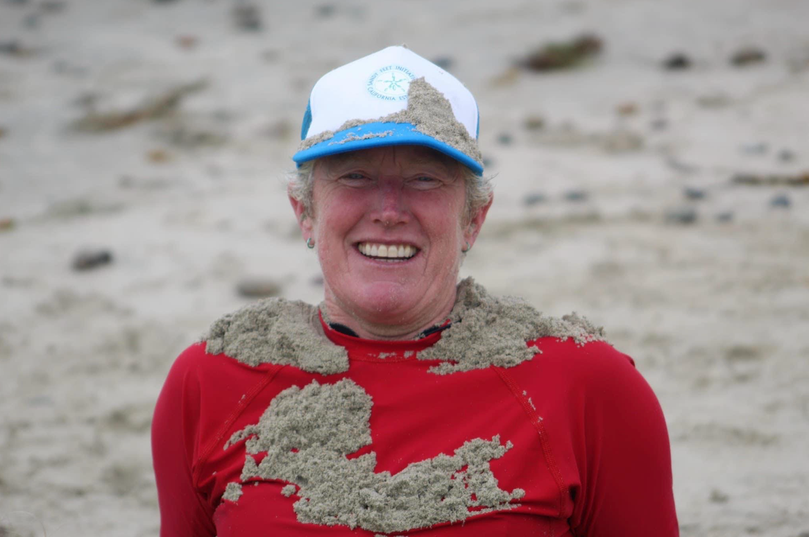 Smiling person in a red shirt and white cap, covered in sand on a beach