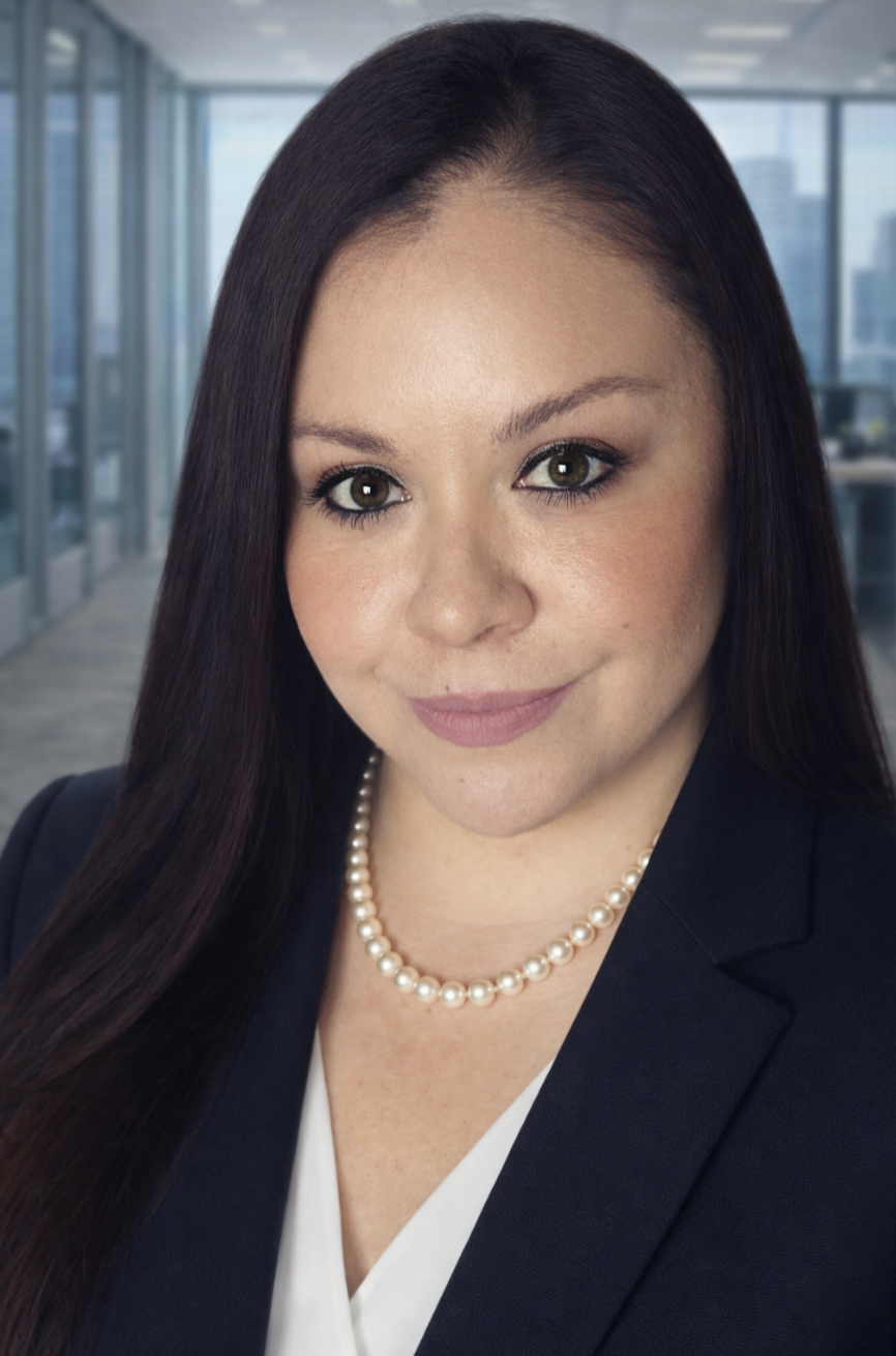 Professional headshot of a woman in a dark blazer and pearl necklace, standing in a blurred office hallway.