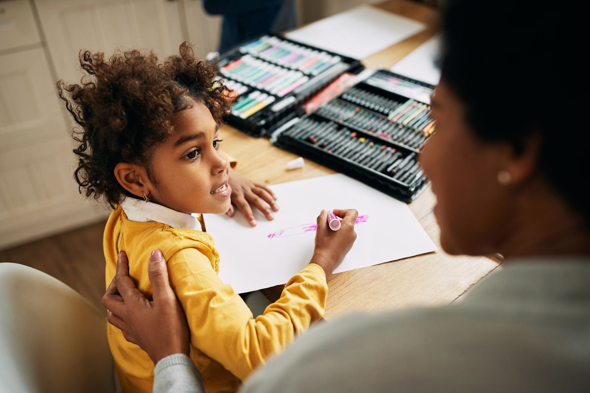 Child drawing at a table while an adult guides them, with art supplies spread nearby.