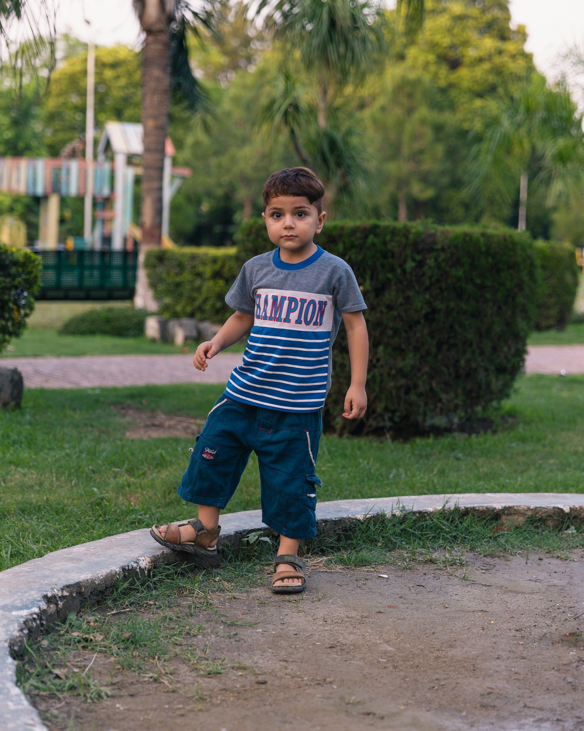 Young child standing on a grassy path in a park, wearing a striped T-shirt and sandals.
