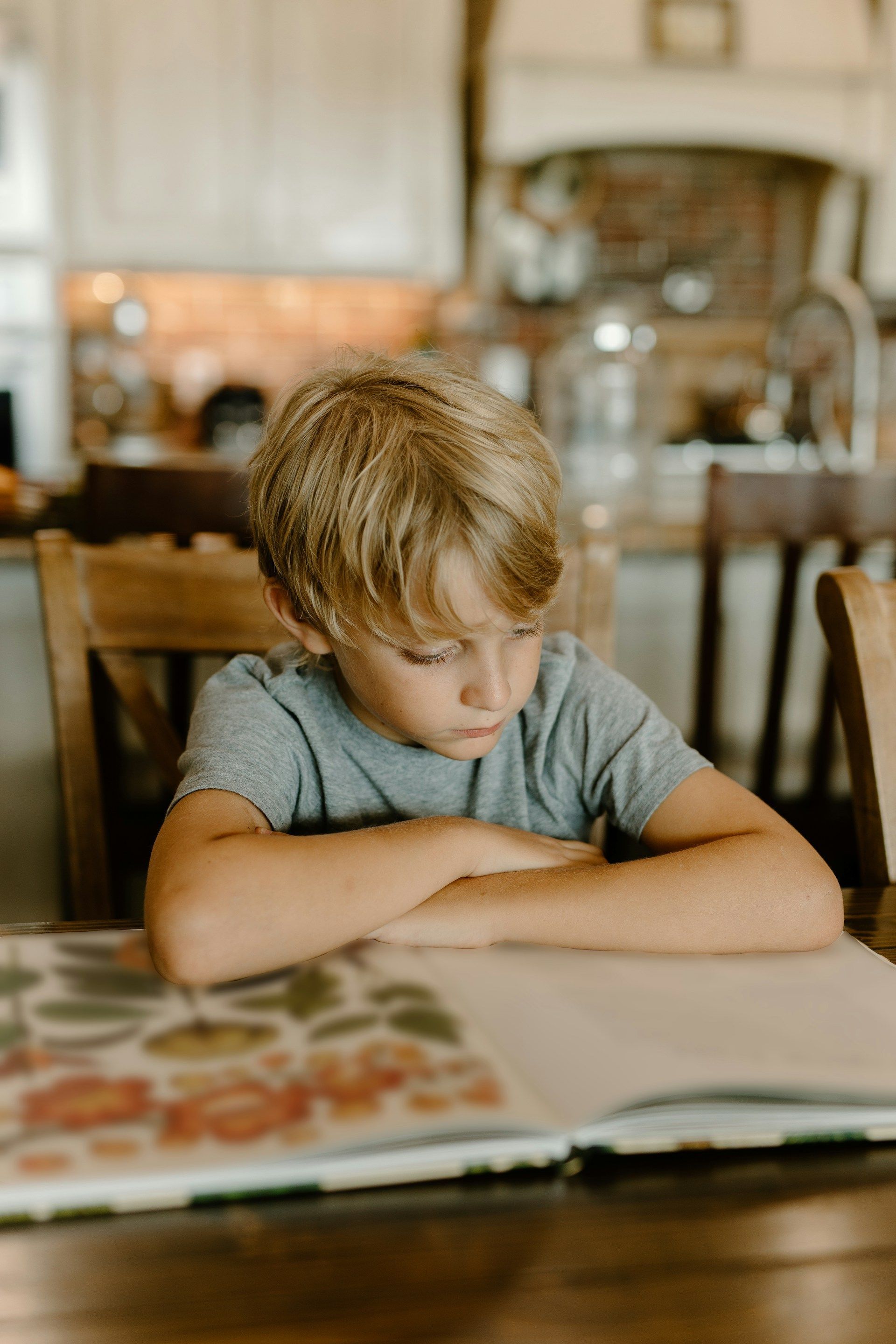 Child reading a book at a kitchen table, leaning on folded arms.