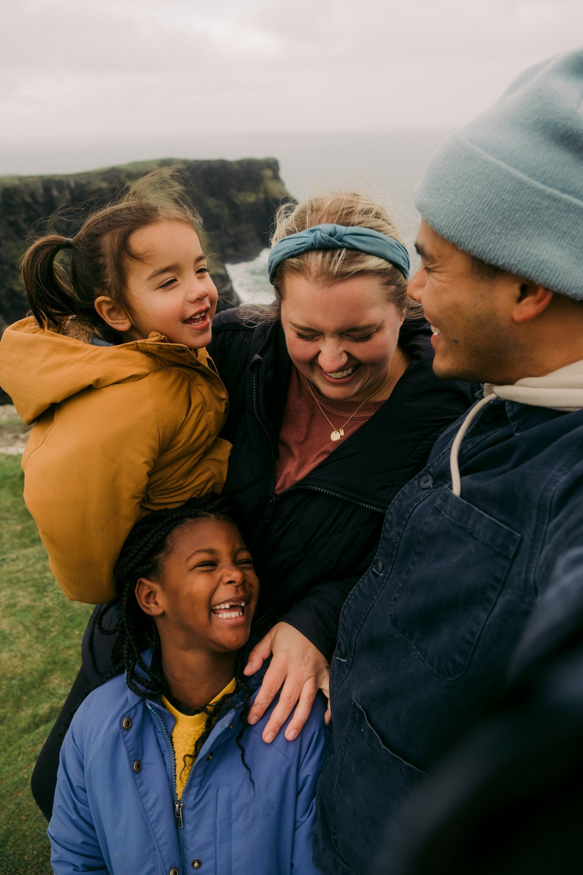 Four people smiling and hugging outdoors by a rocky shoreline in cold weather.