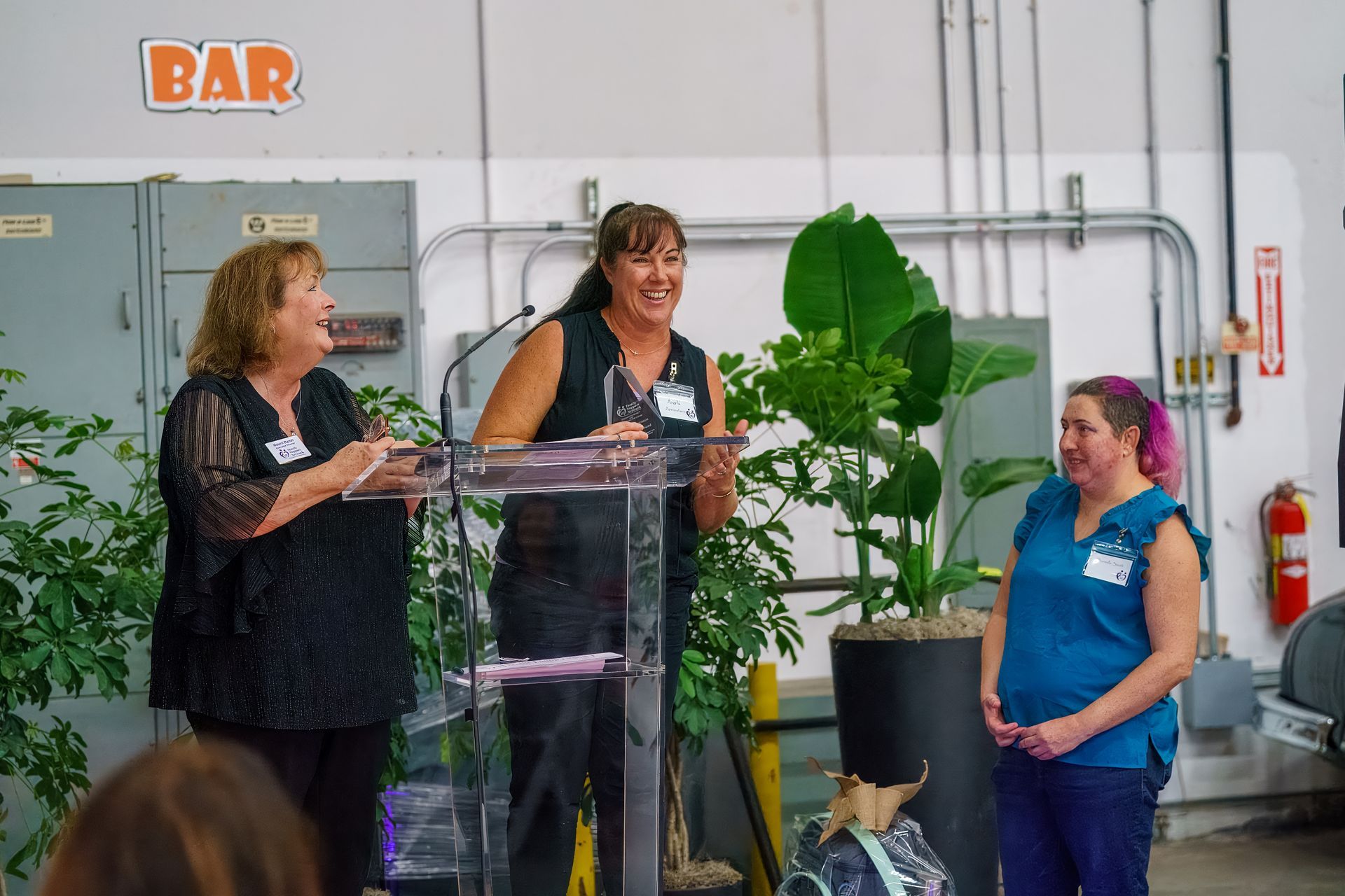 Three people at a podium in a warehouse, with a BAR sign and a large potted plant nearby.