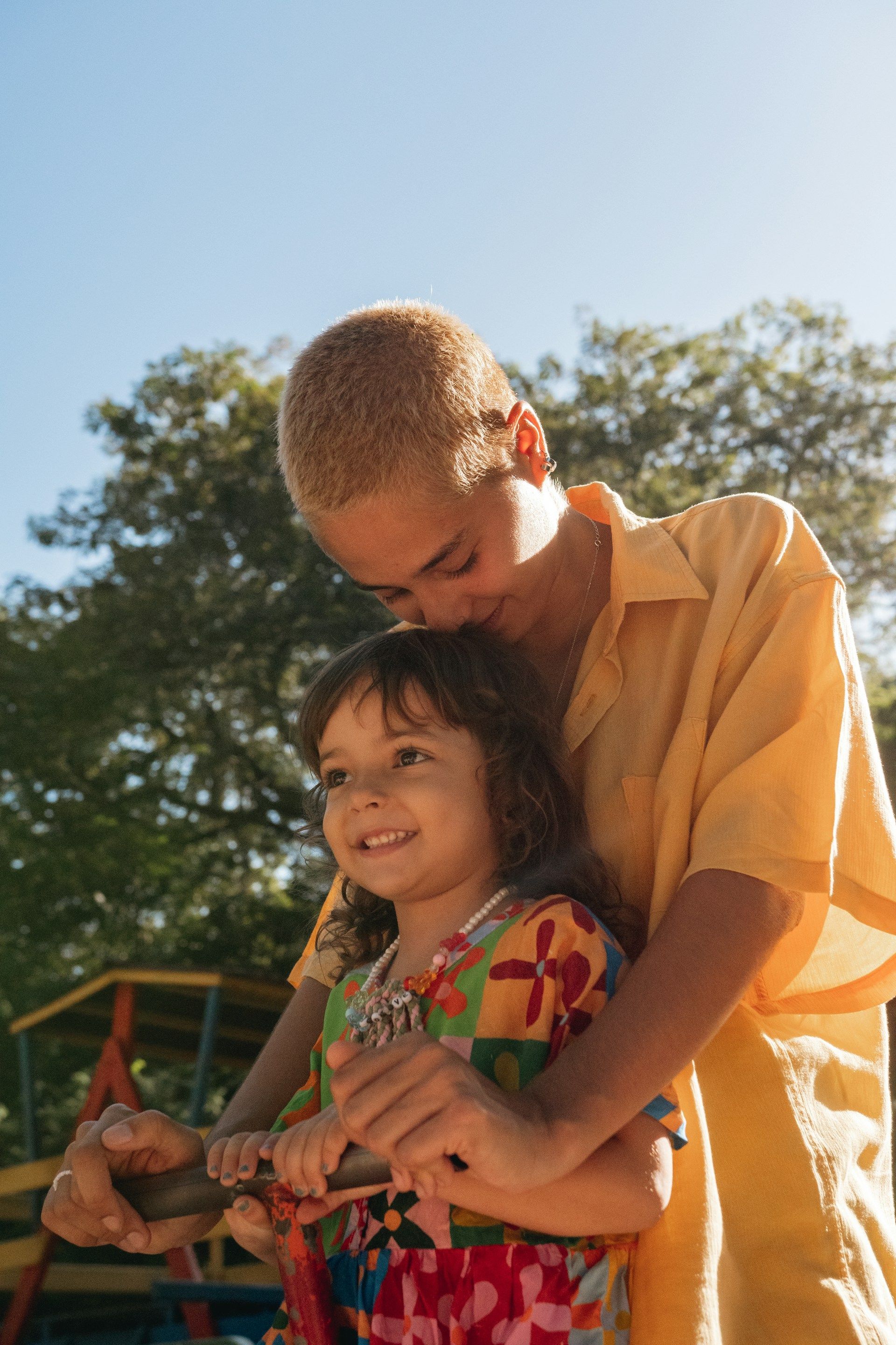 Two children hugging outdoors, smiling in warm sunlight with trees in the background