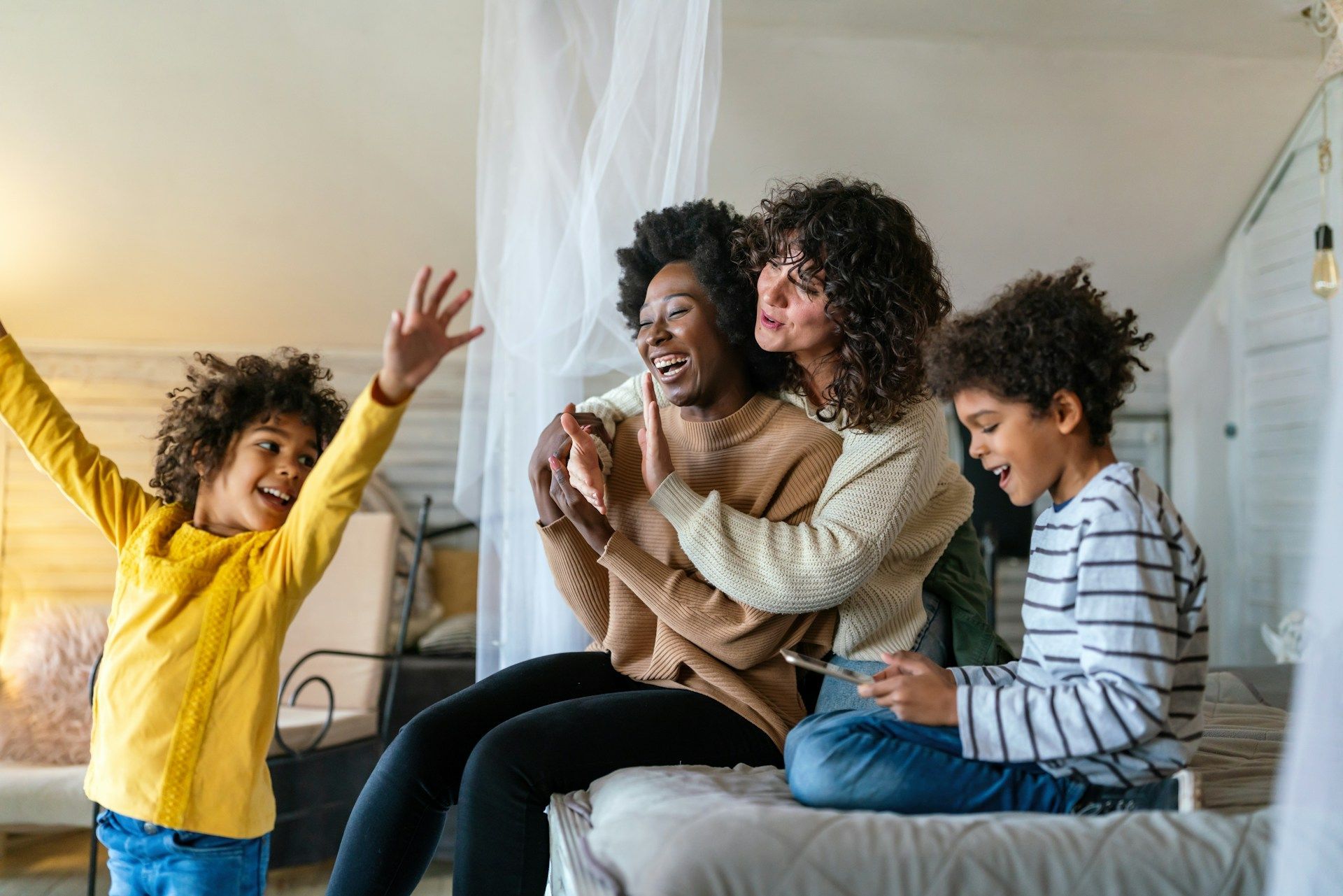Family sitting on a bed, smiling and playing together in a bright bedroom.
