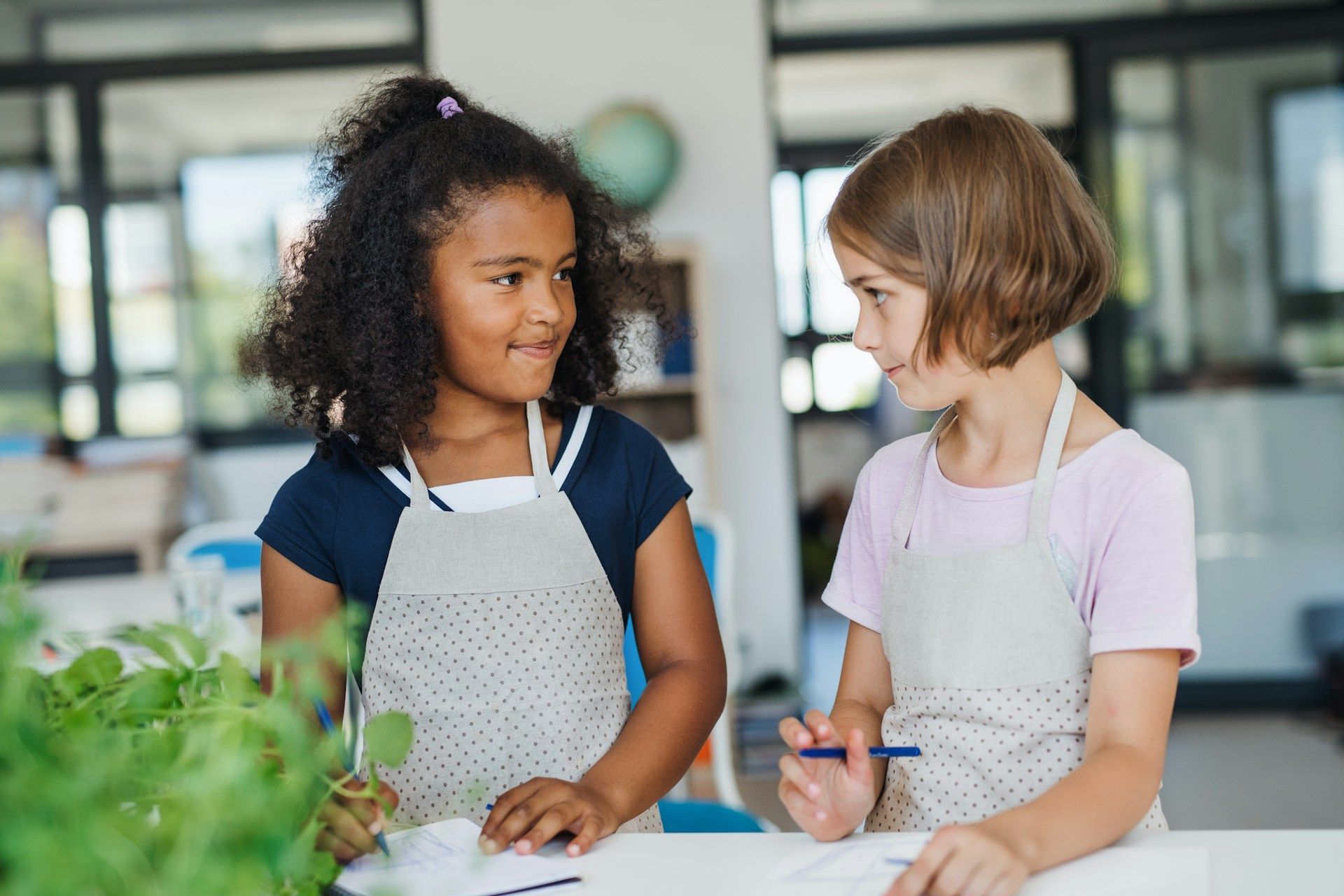 Two children in aprons preparing food together at a kitchen counter, smiling and talking.