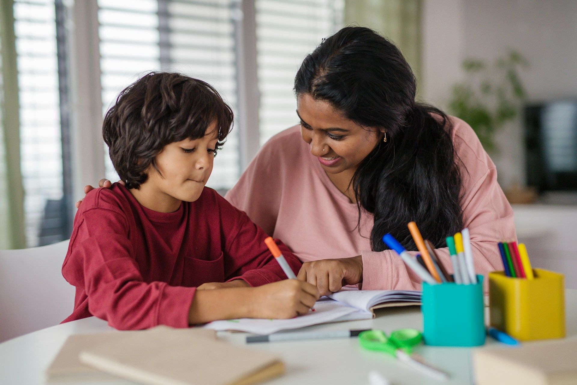 Adult and child writing together at a table with notebooks and colorful pencils