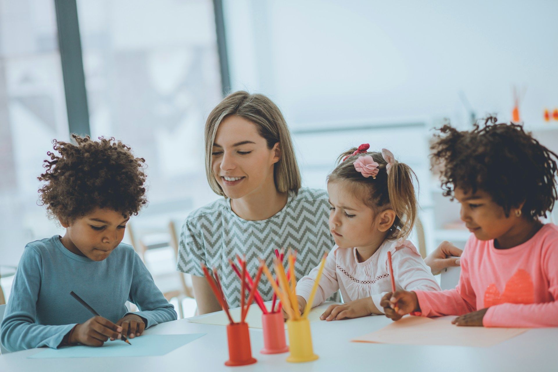 Teacher and three children making crafts at a table with colorful crayons in a bright classroom