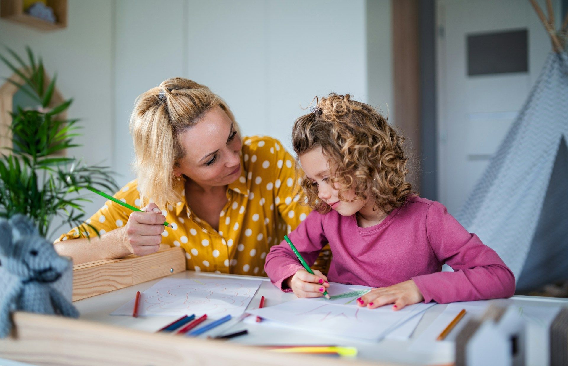 Woman and child drawing together at a table, smiling in a bright room.