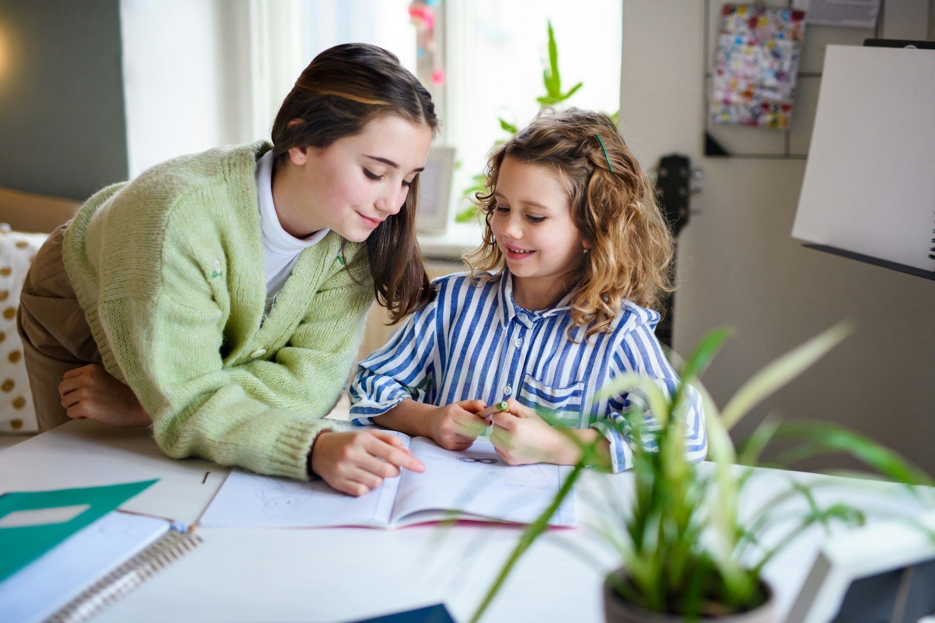 Teacher helping a child write at a table with open notebooks and a plant in the foreground