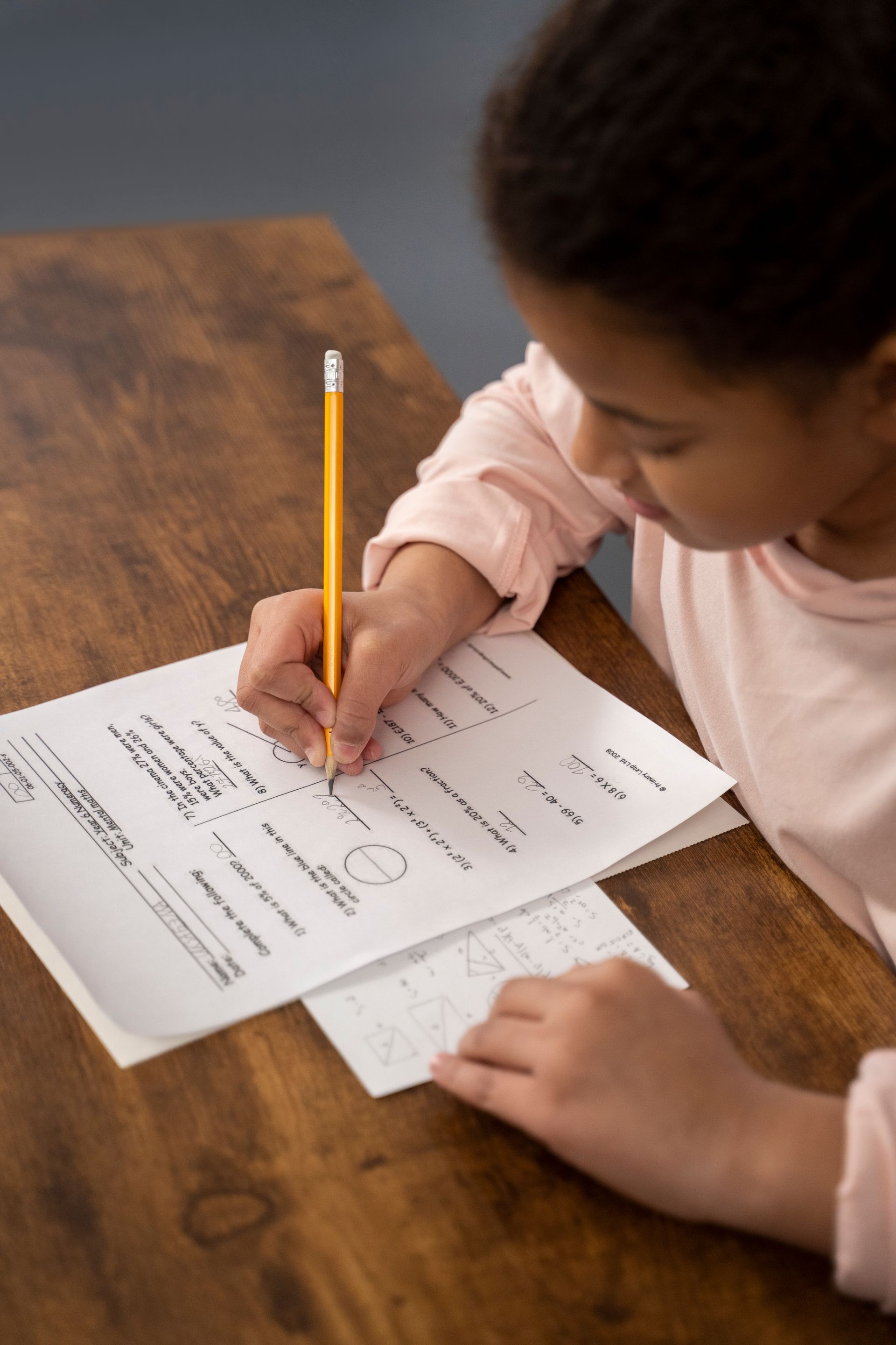 Child writing on a worksheet at a wooden table with a pencil.