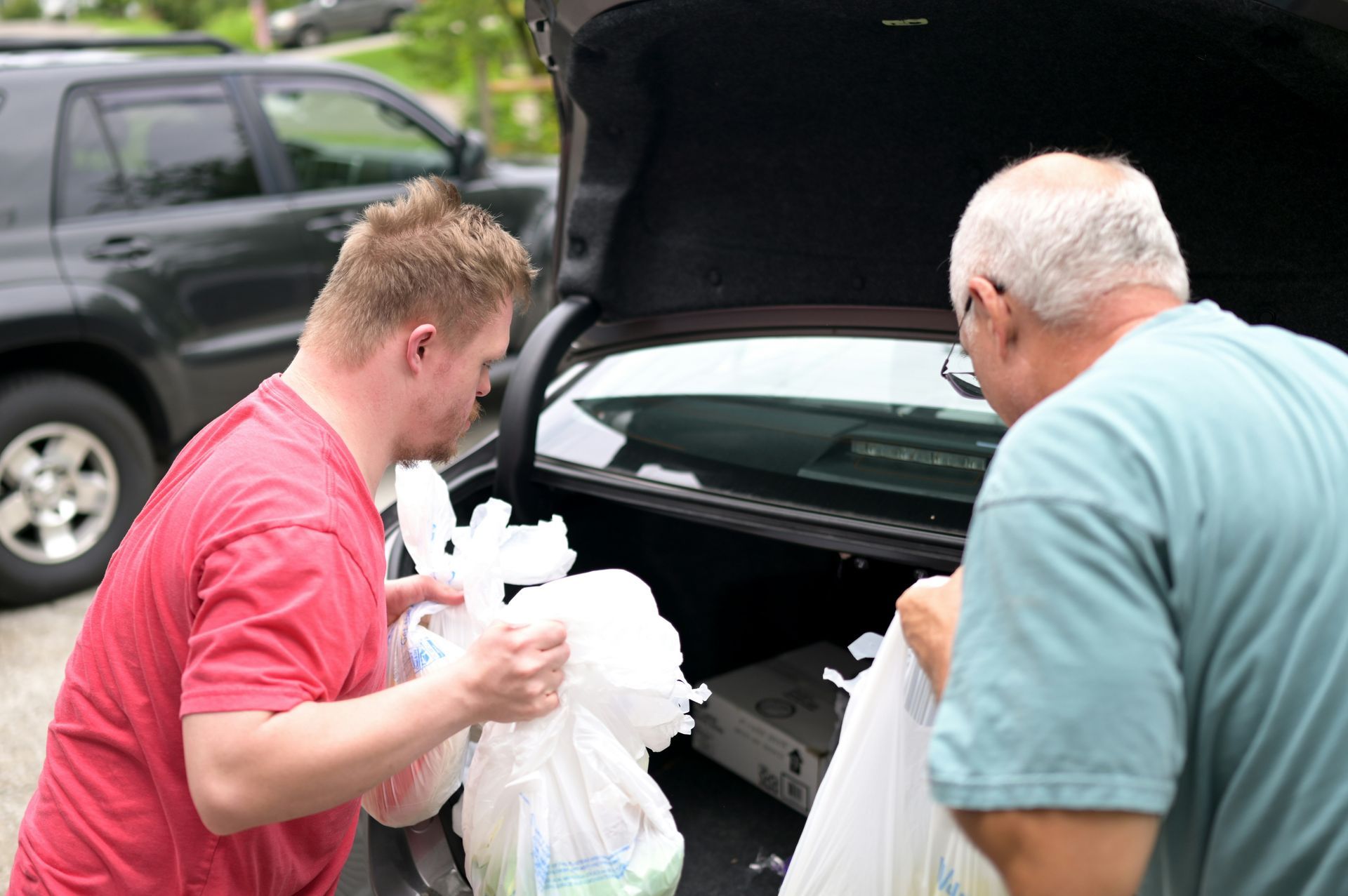 Two people load white bags into the open trunk of a car outdoors