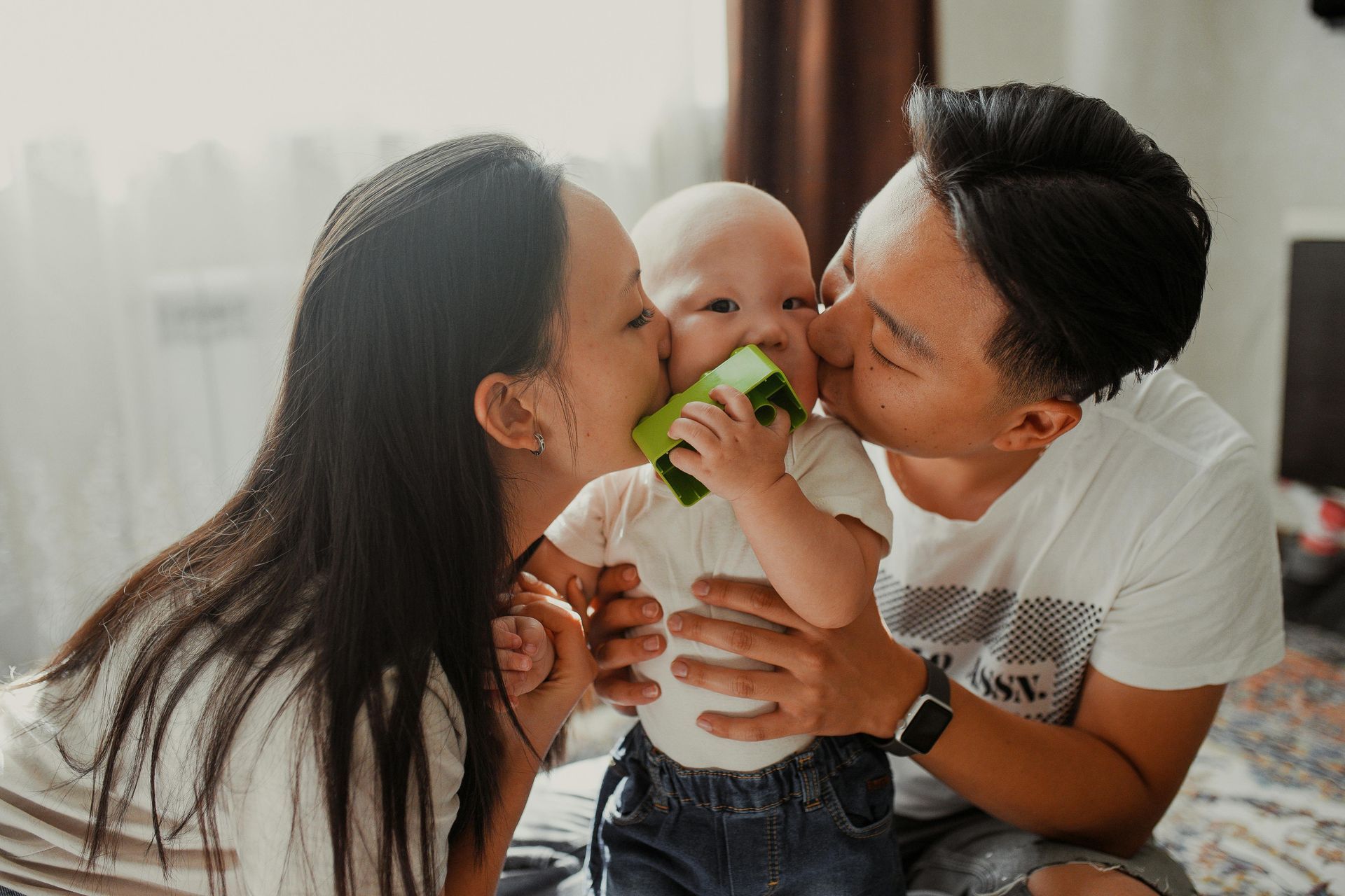 Parents kissing baby while holding a green toy, indoors near a bright window