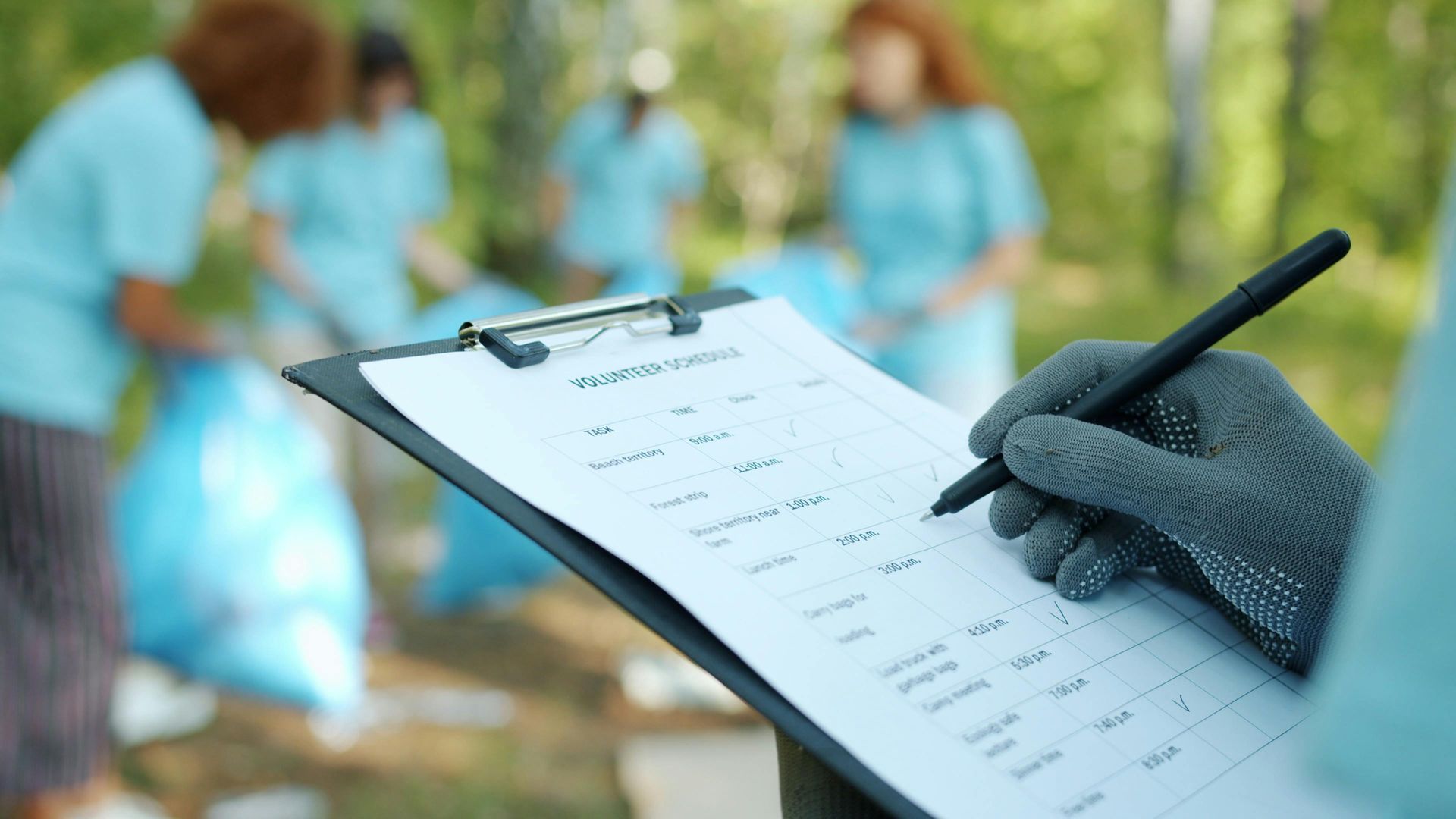 Gloved hand holding a clipboard checklist in a garden with blurred volunteers in blue uniforms