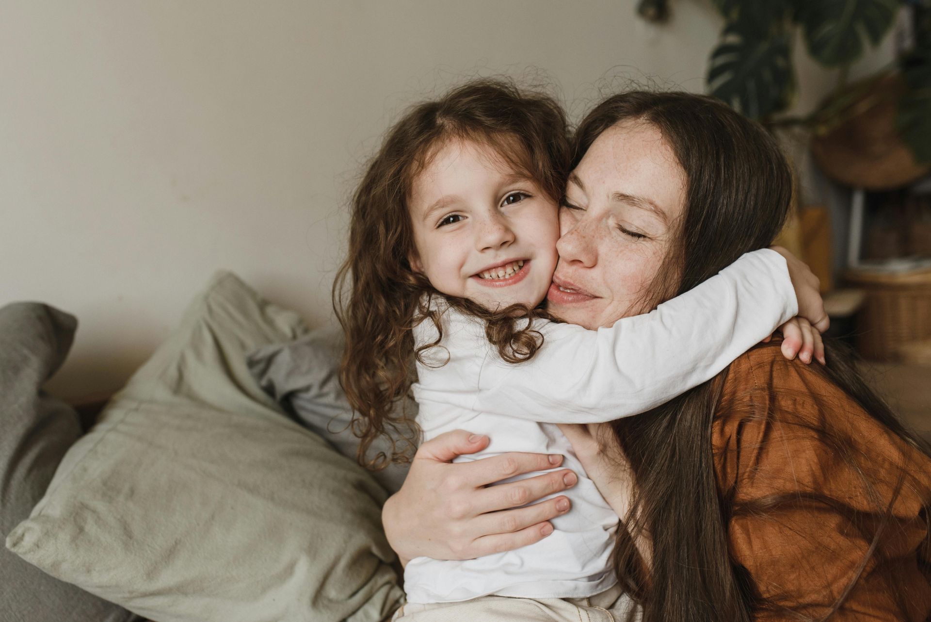 Woman cuddling a smiling child on a couch, both embracing warmly indoors.