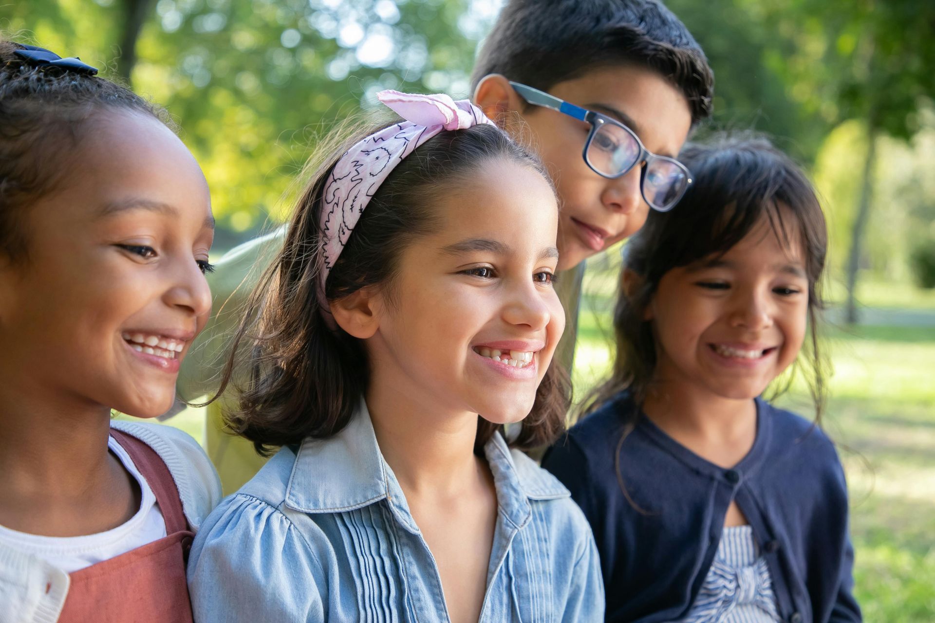 Four smiling children standing outdoors in a sunny park, with trees in the background.